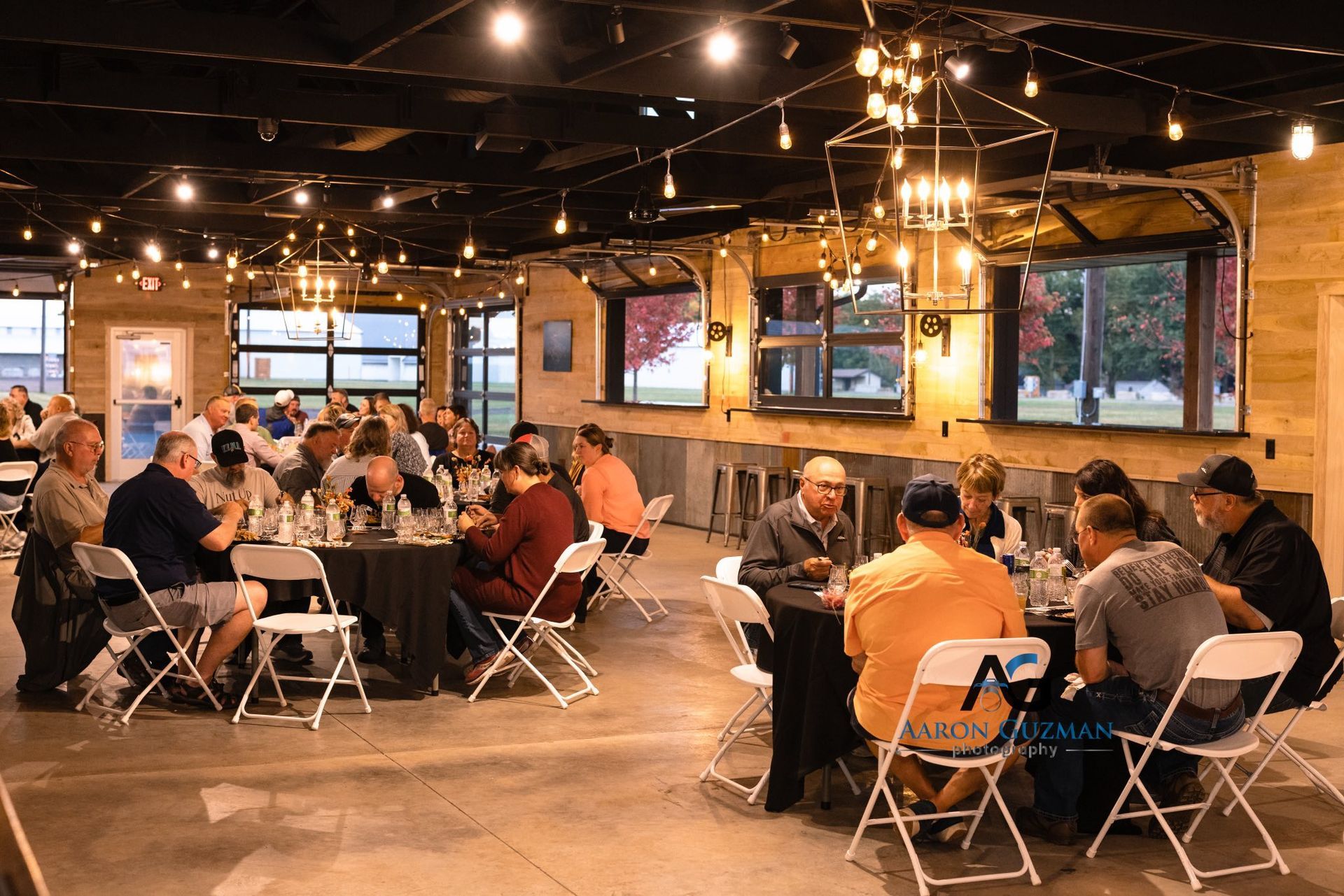 People sitting at tables in a brightly lit, rustic indoor event space with wooden walls and large glass doors.