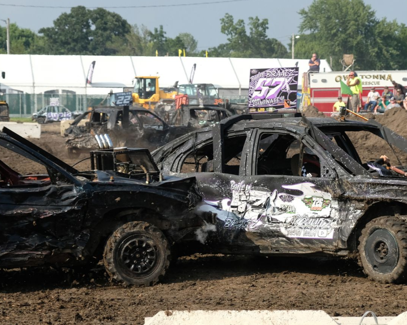 Two crushed race cars collide in a muddy demolition derby arena, with spectators in the background.