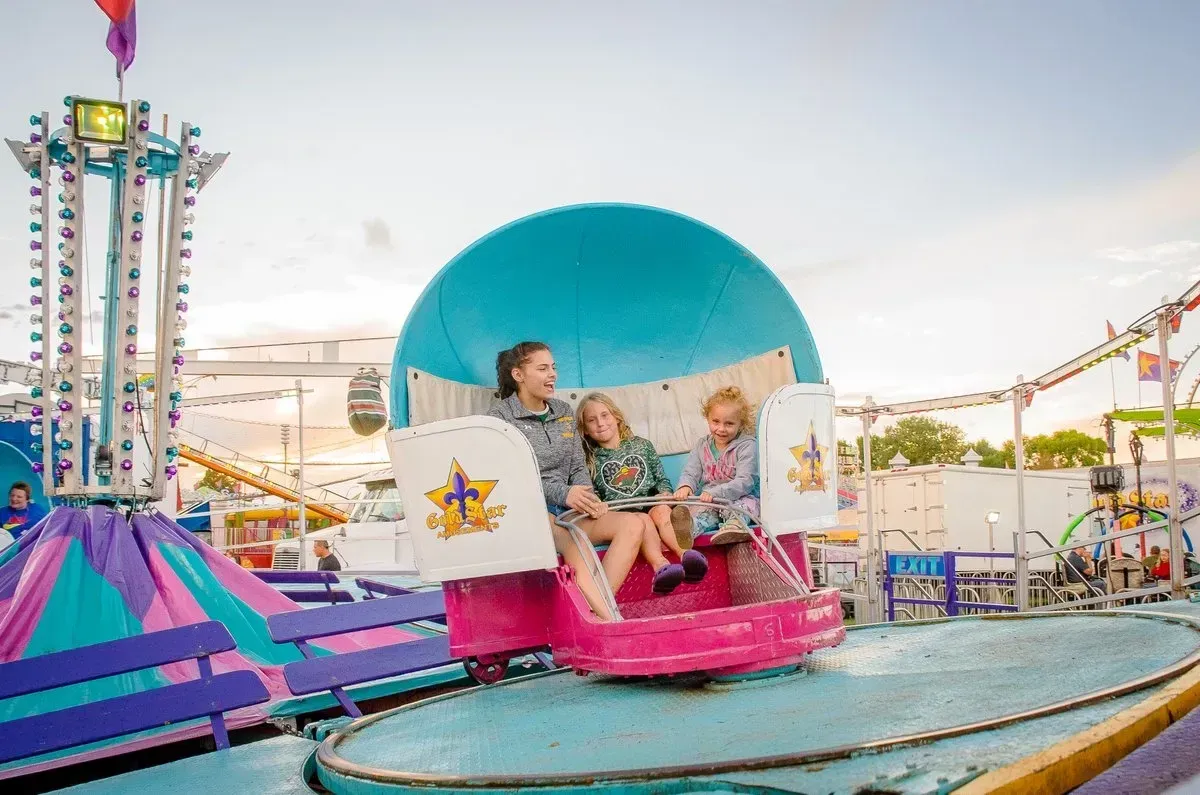 Three people ride a pink amusement park ride with a blue sphere backdrop. They are smiling. Other rides are visible.