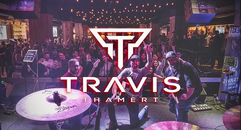 Crowded concert venue with a large Travis logo and name over a purple-lit stage and audience