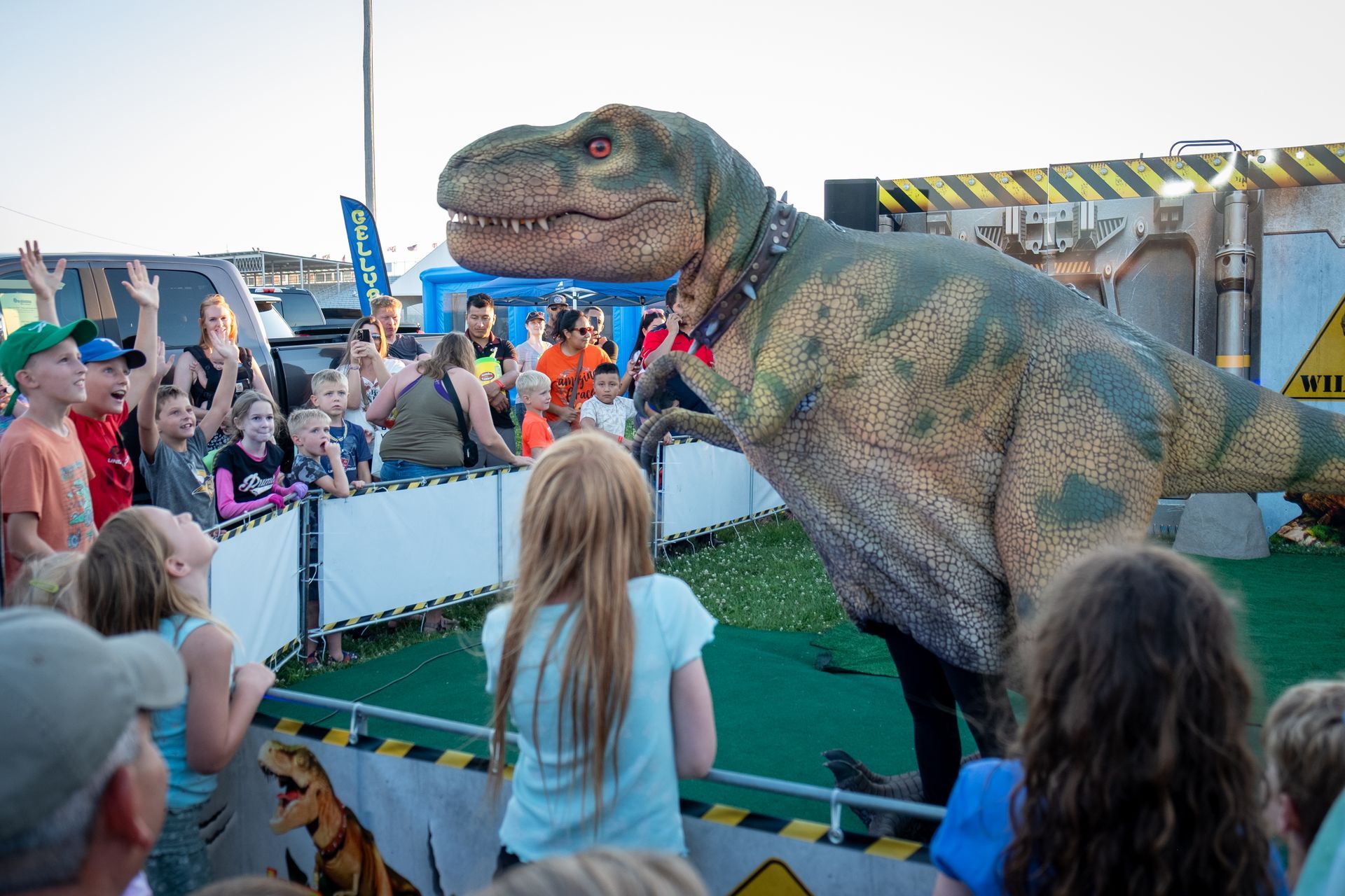 Crowd watches a life-sized T. rex puppet at an outdoor event behind a barrier.