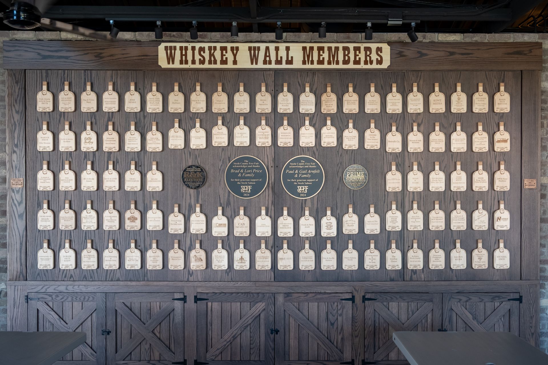 Whiskey Wall Members: Wooden display of numerous bottle-shaped plaques with names, above a cabinet with doors.