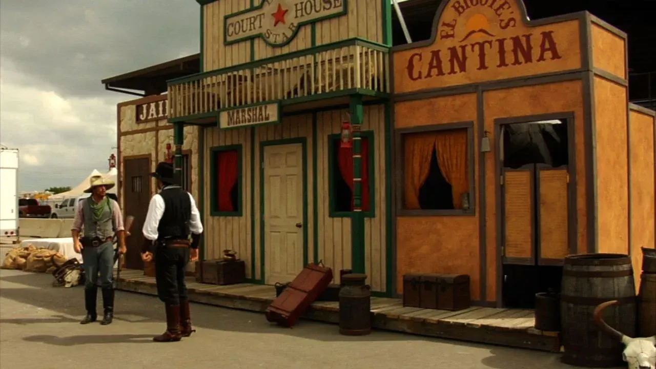 Two men stand outside a rustic cantina storefront with wooden walls and a porch in a dusty western town.