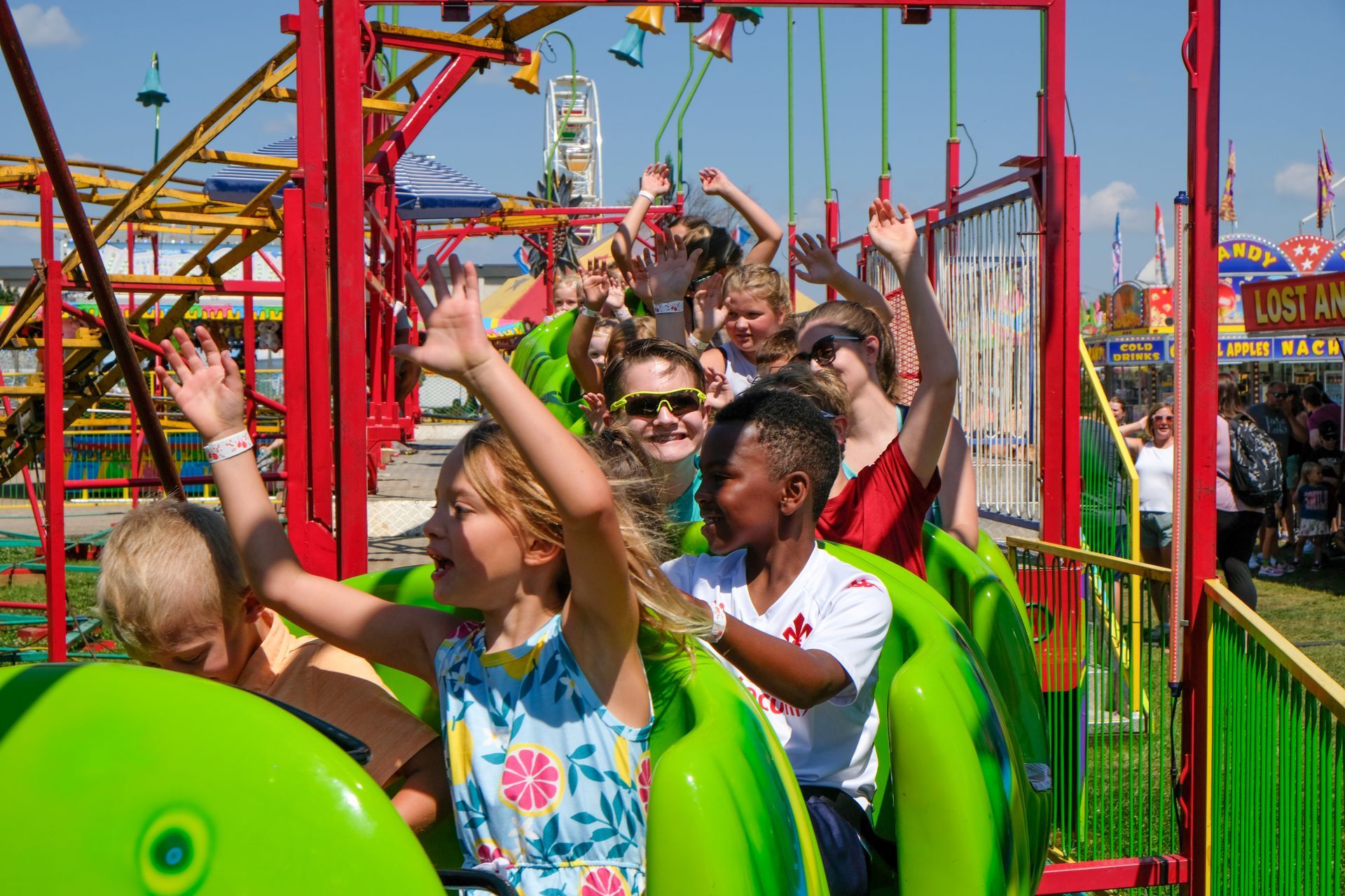 Children on a small roller coaster, waving with joyful expressions, at a brightly-colored amusement park.