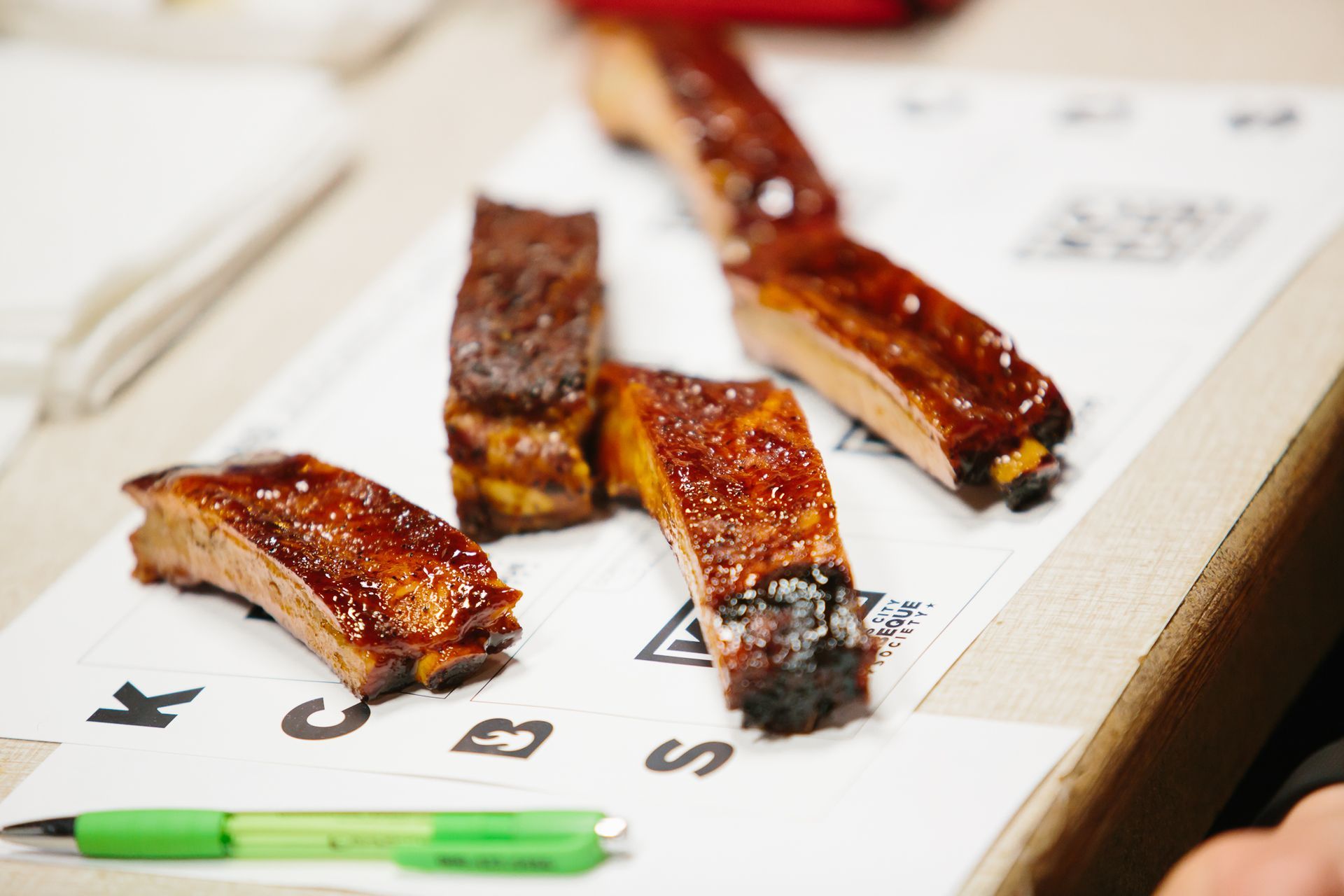A display of glazed barbecue ribs on a white tray with a green pen beside it.