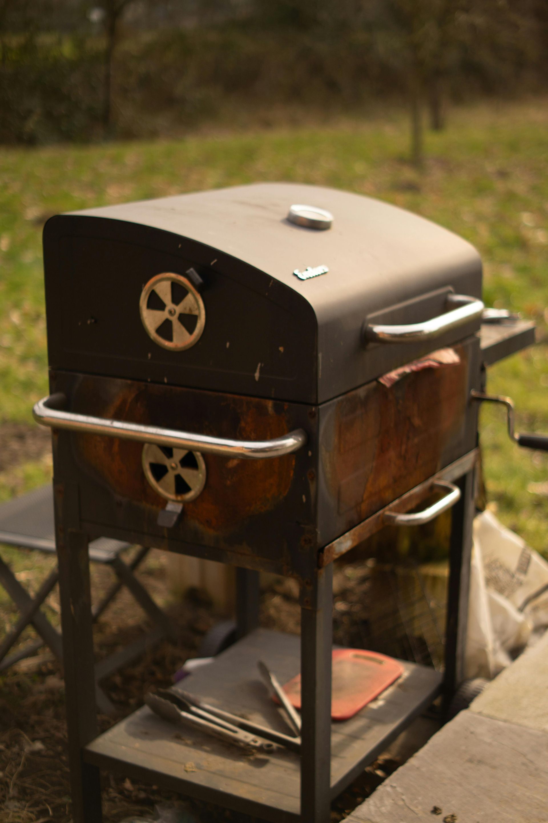 Metal grill or smoker outdoors with a black lid and silver handles on a stand.