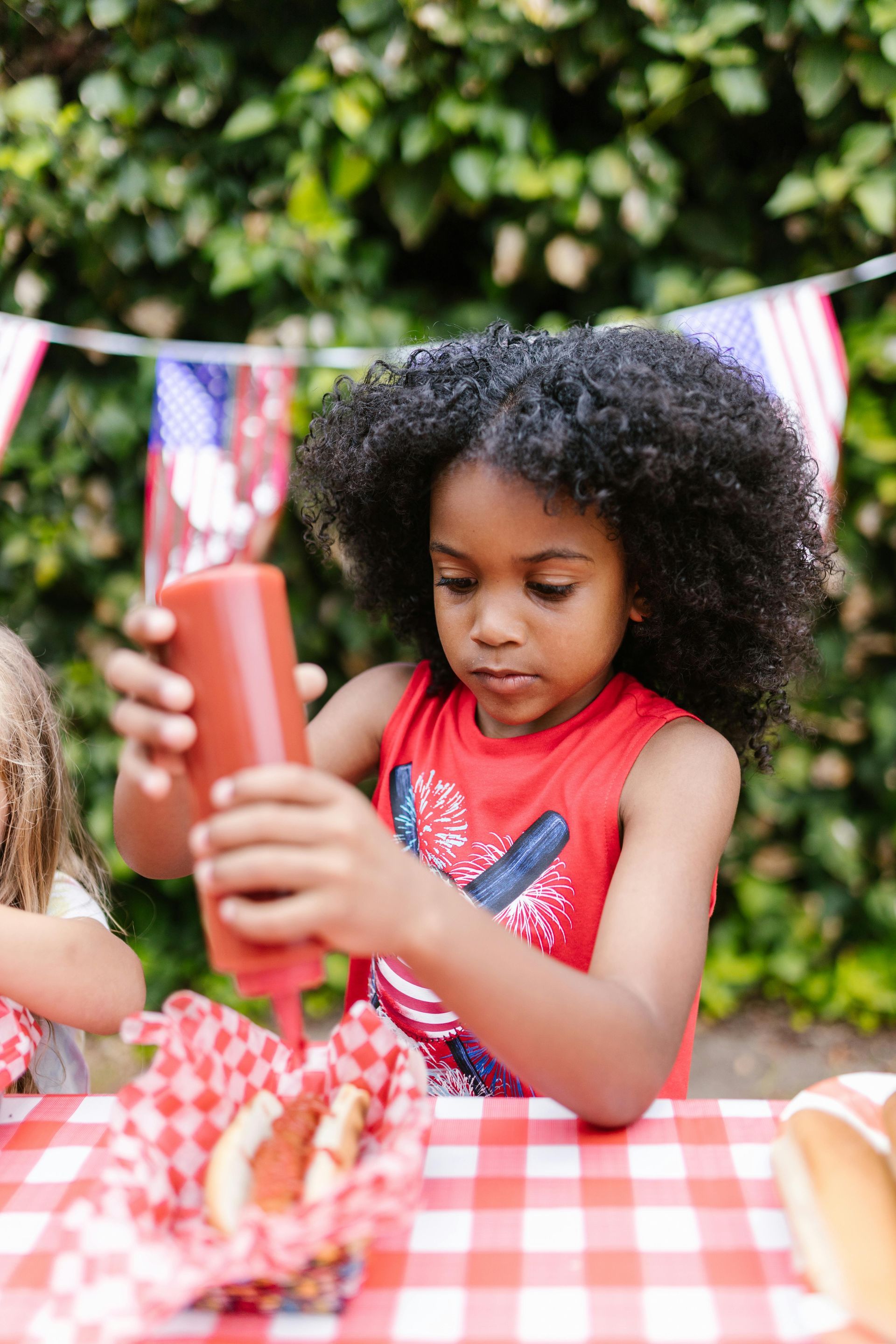 Child at a picnic table holding a red cup, with a checkered tablecloth and flags in the background