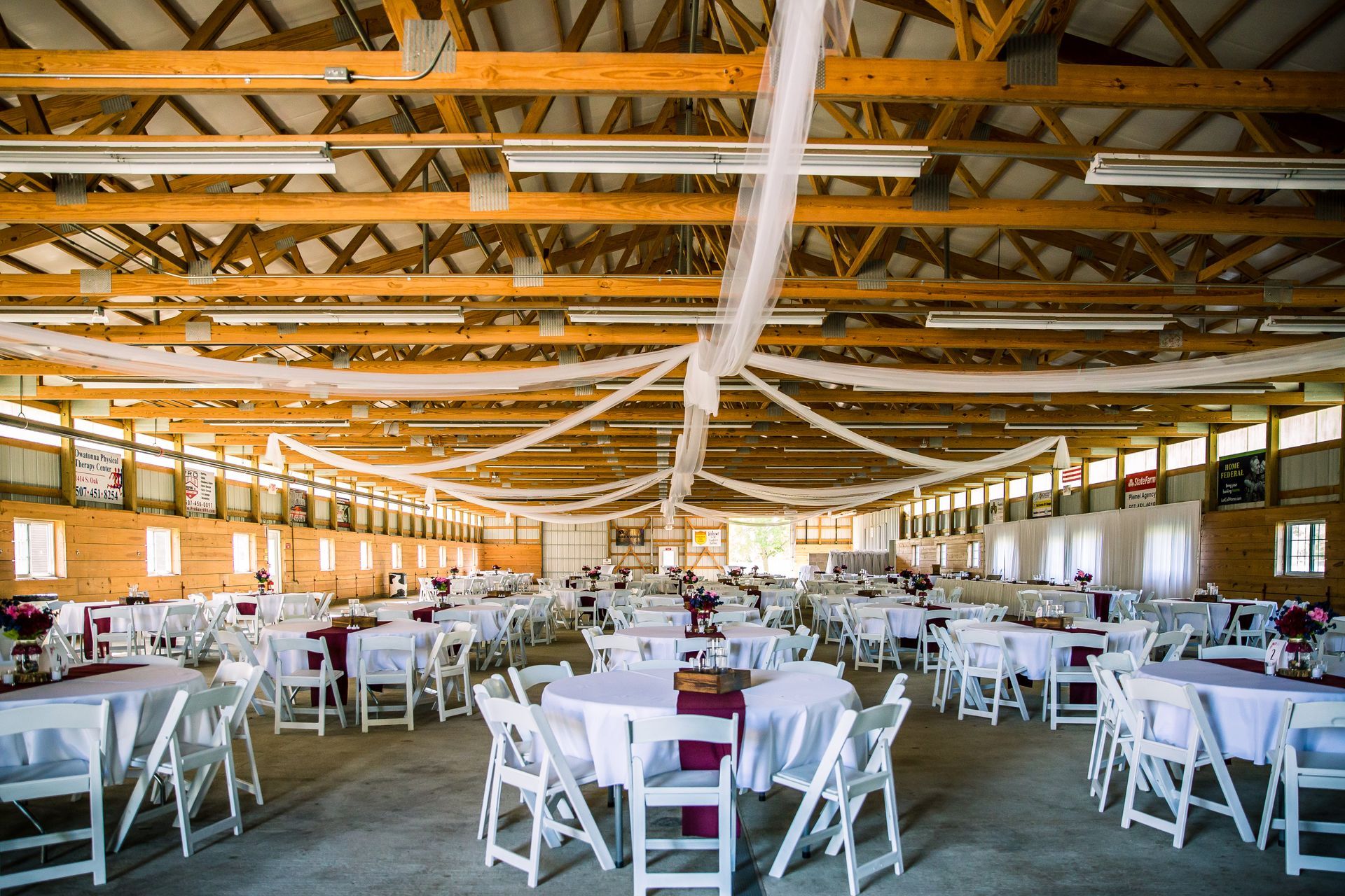 A large, rustic event barn interior set up with round dining tables, white chairs, and elegant white fabric ceiling drapes.