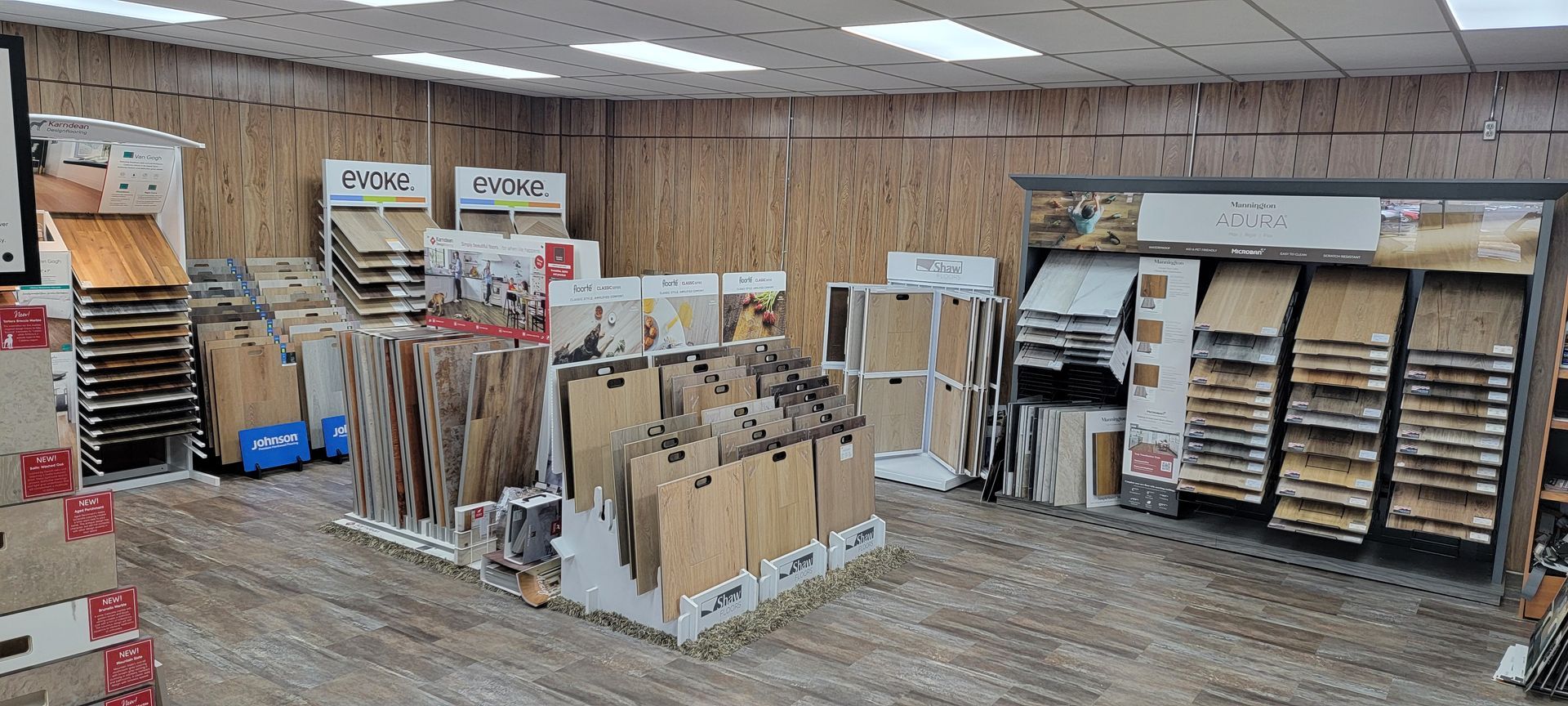 A flooring store interior with displays of wood flooring samples. Wood-paneled room with stacked shelves, racks of hanging samples, and rolled materials on display