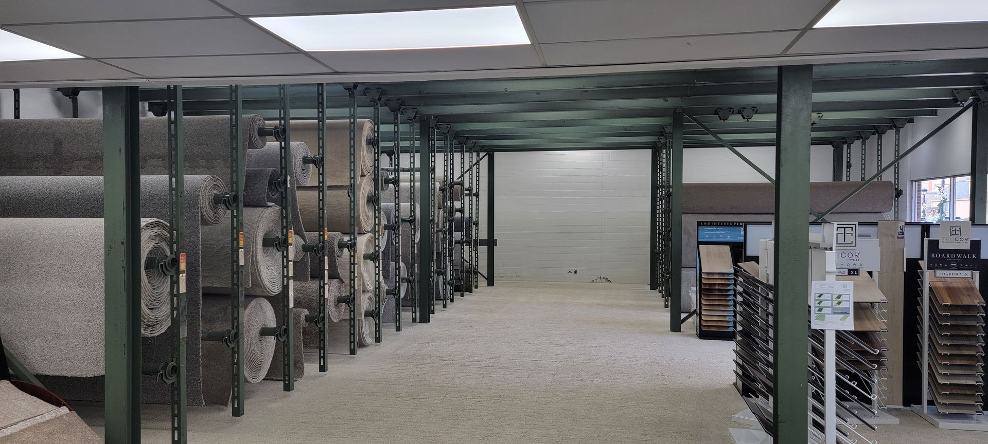 Interior of a carpet store with rolls of carpet on racks and a person working. Storage room with metal shelving, stacked boxes, and a carpeted aisle under fluorescent lights
