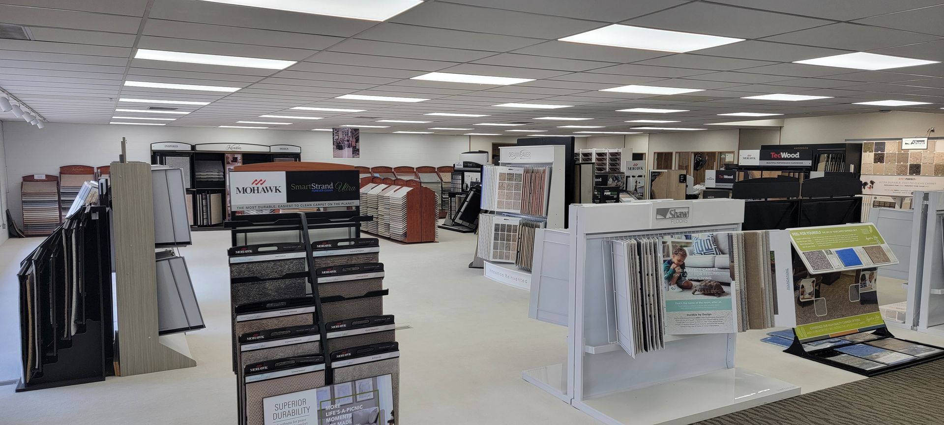 A retail showroom with flooring and display materials on tables and racks under bright overhead lights. Bright retail showroom with fabric, wallpaper, and decor sample displays on white stands