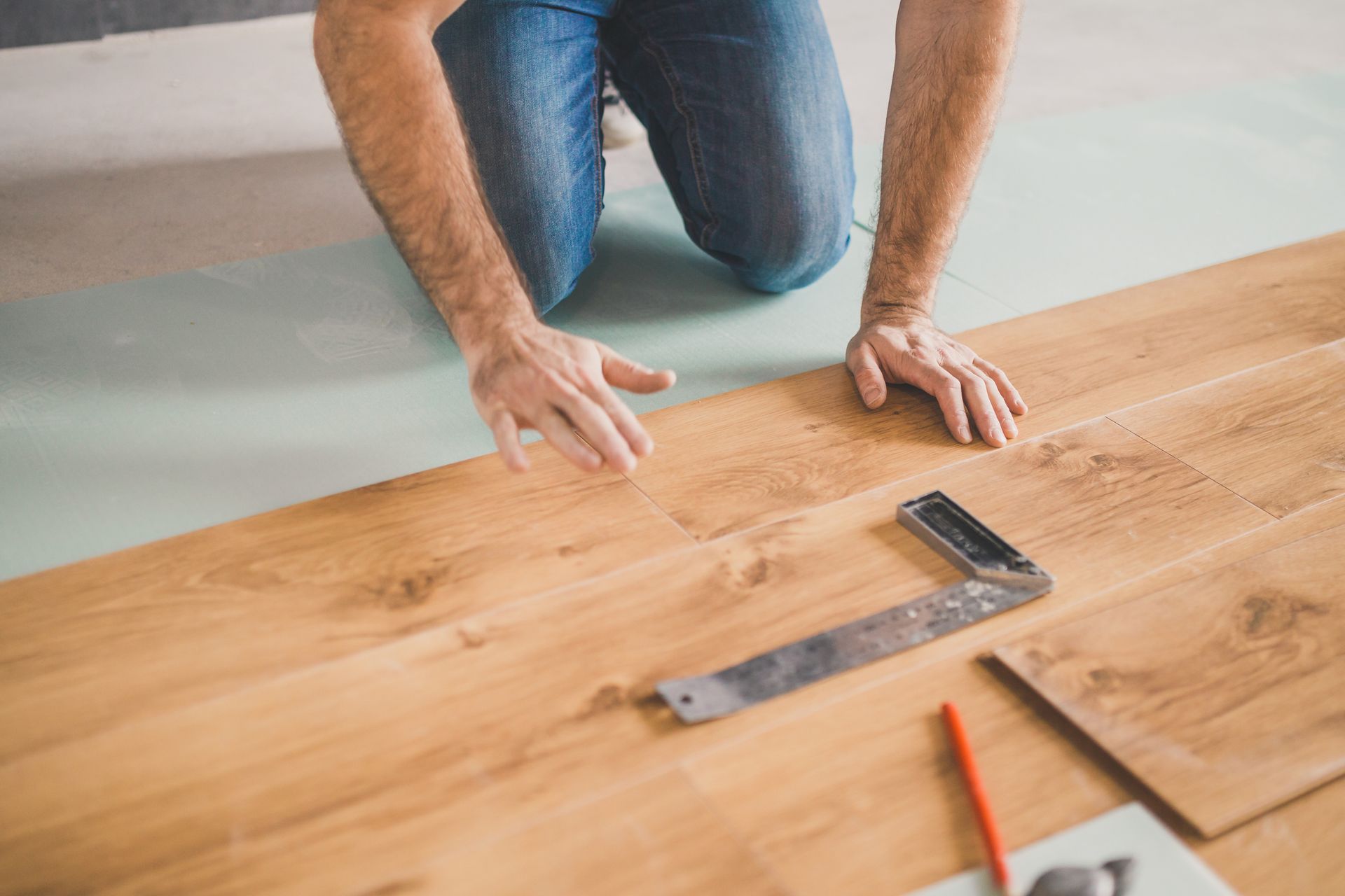 Person kneeling on a wooden floor, measuring a plank with a ruler and pencil to install flooring