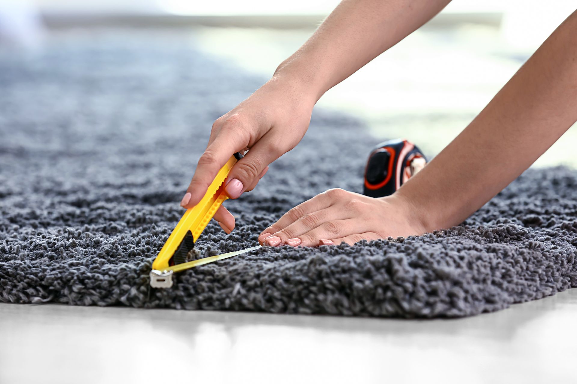 Woman with tape measure cutting carpet on floor. Hands using a yellow utility knife to cut a dark gray carpet on a white floor.