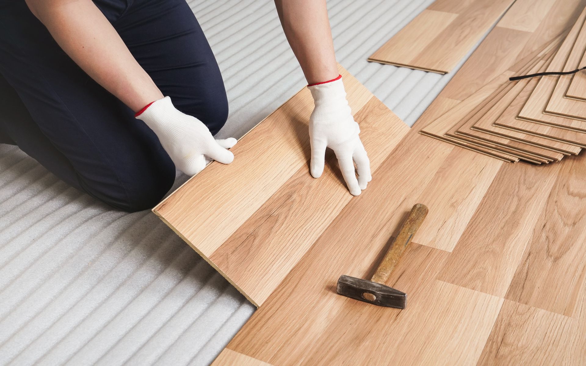 Person installing laminate flooring, wearing gloves, with a hammer nearby. Hands installing light wood flooring with a hammer and spacers on a subfloor.
