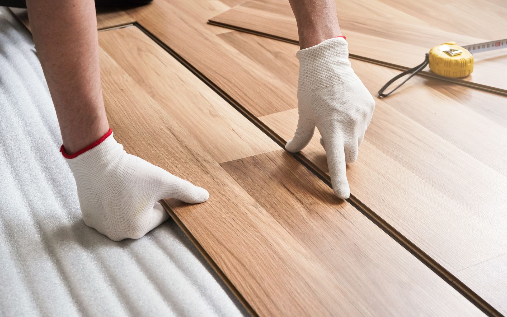 Hands in white gloves installing wood floor planks. A measuring tape is nearby. Gloved hands measuring and laying wooden floorboards with a tape measure nearby