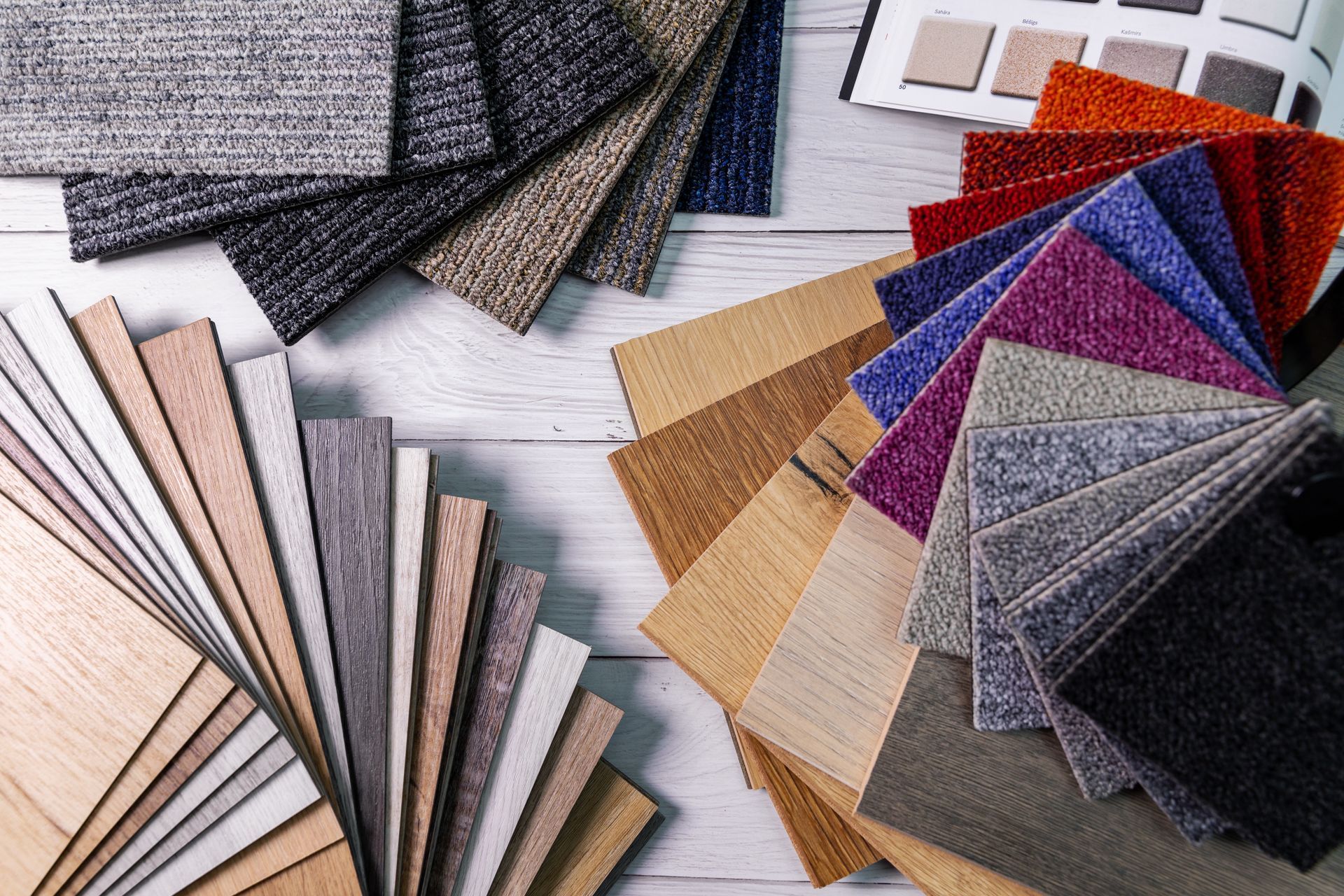 Assortment of flooring samples including wood, carpet, and textile swatches in various colors, arranged on a light wood surface. Assorted fabric swatches and sample books in neutral, gray, beige, red, and blue tones on a wooden surface