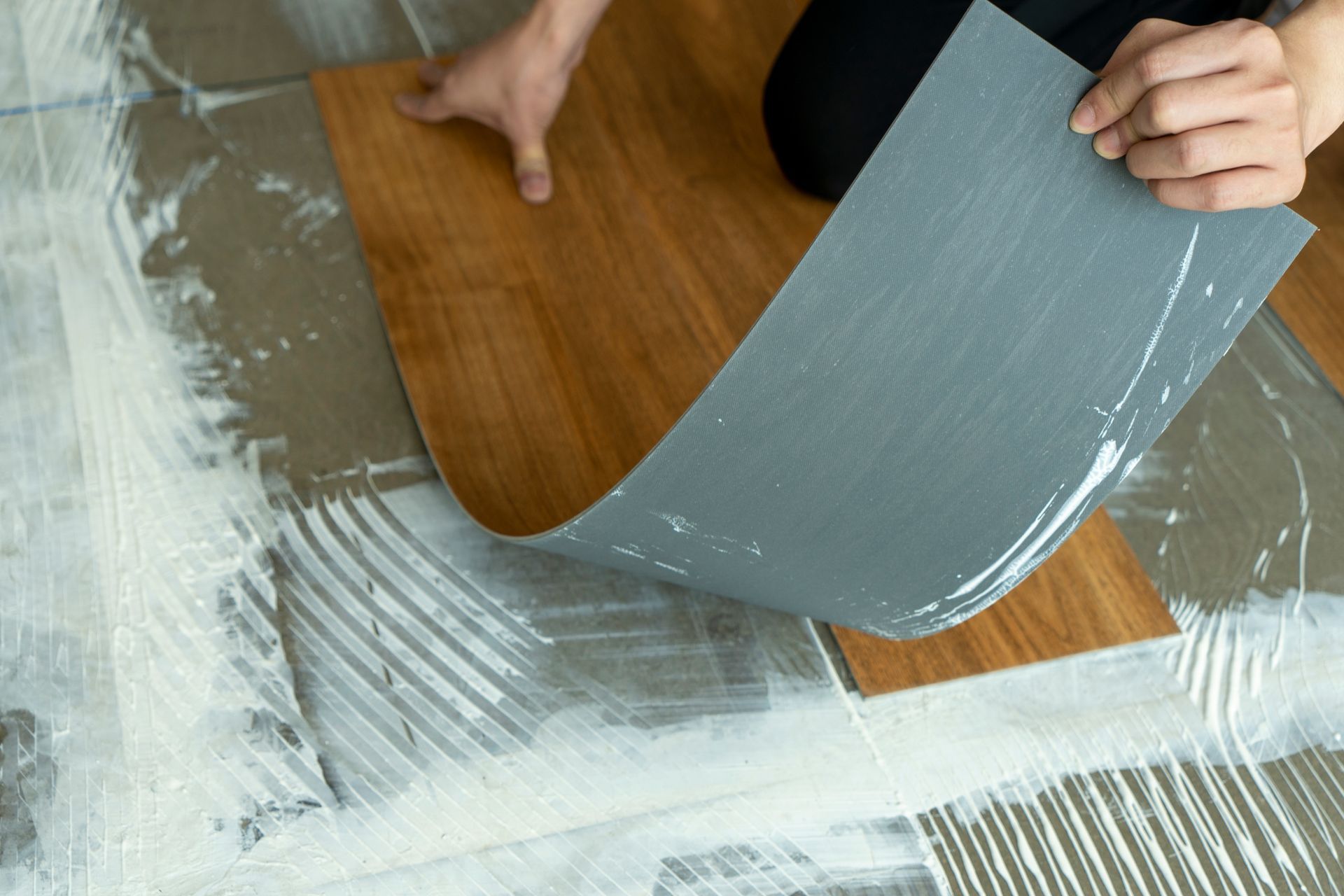 Hands spread gray adhesive on a floor over white underlayment with a trowel.