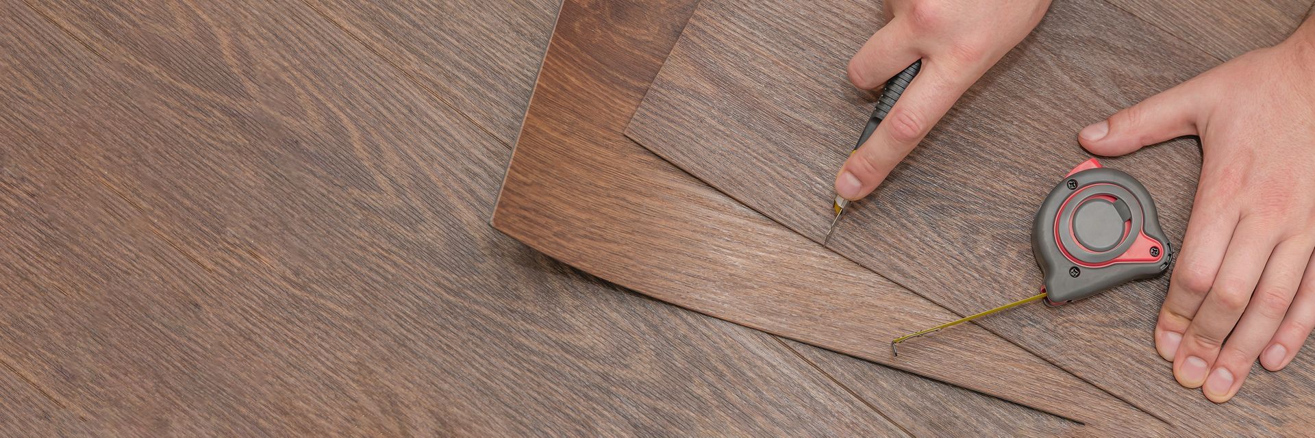 Hands using a tape measure to measure brown fabric on a worktable.