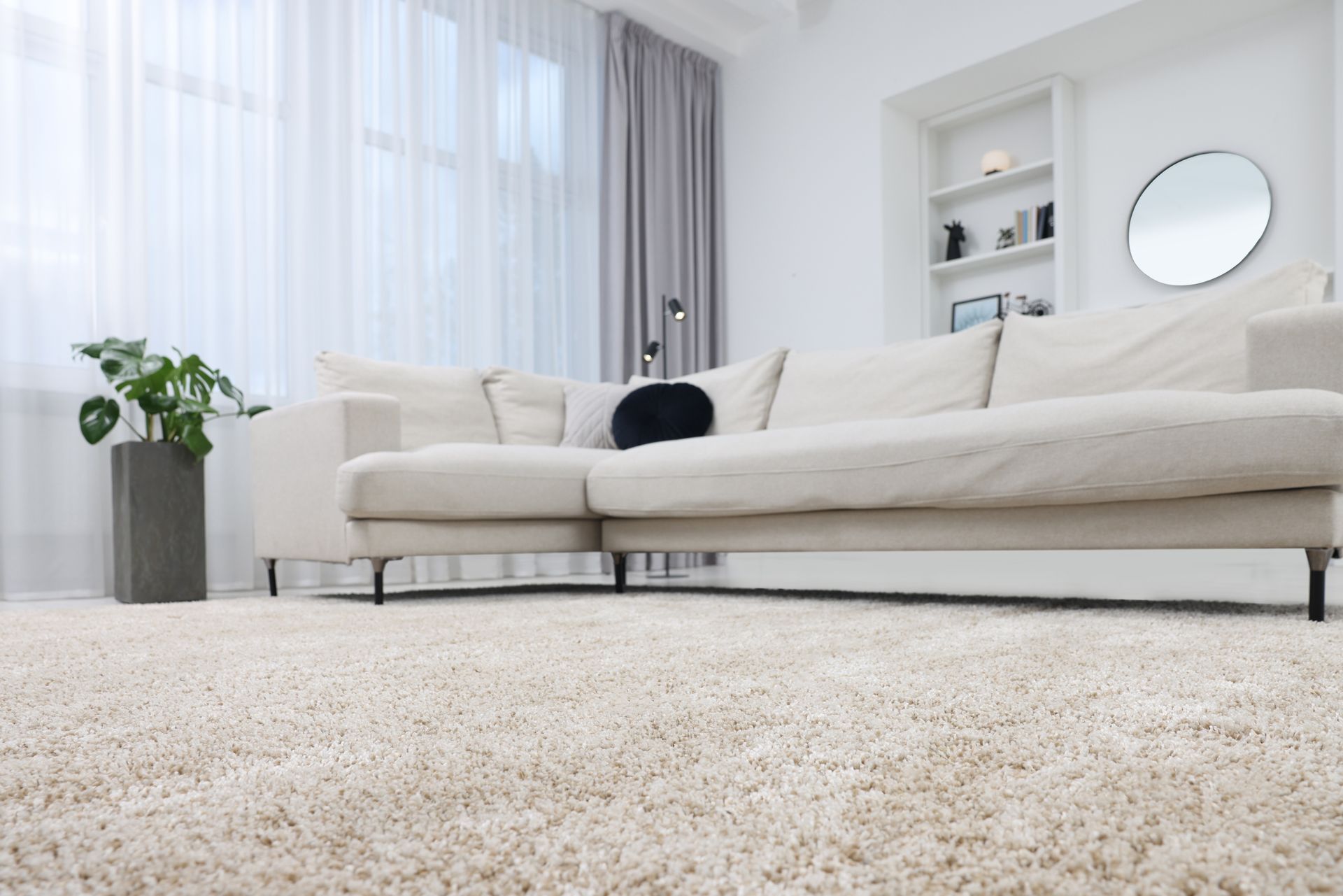 Living room with a large cream-colored sectional sofa, fluffy rug, and sheer curtains. Bright minimalist living room with a beige sofa, white shelves, sheer curtains, and a textured cream rug.