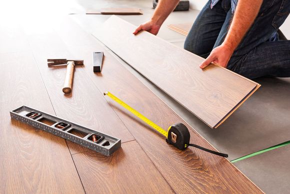Person measuring a laminate floor plank beside tools, tape measure, and level on a wooden floor