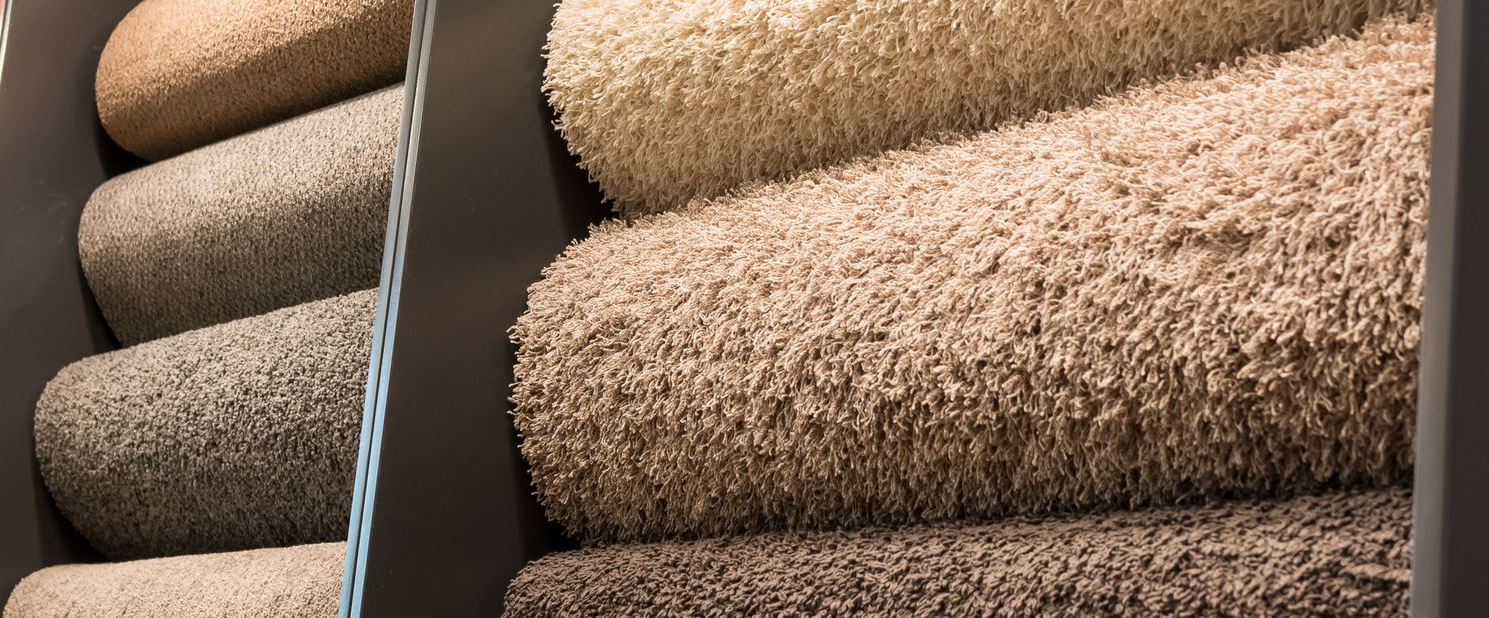 Living room with white sofa, fireplace with logs, large window, and beige carpet. Close-up of beige and brown textured carpets rolled on display racks