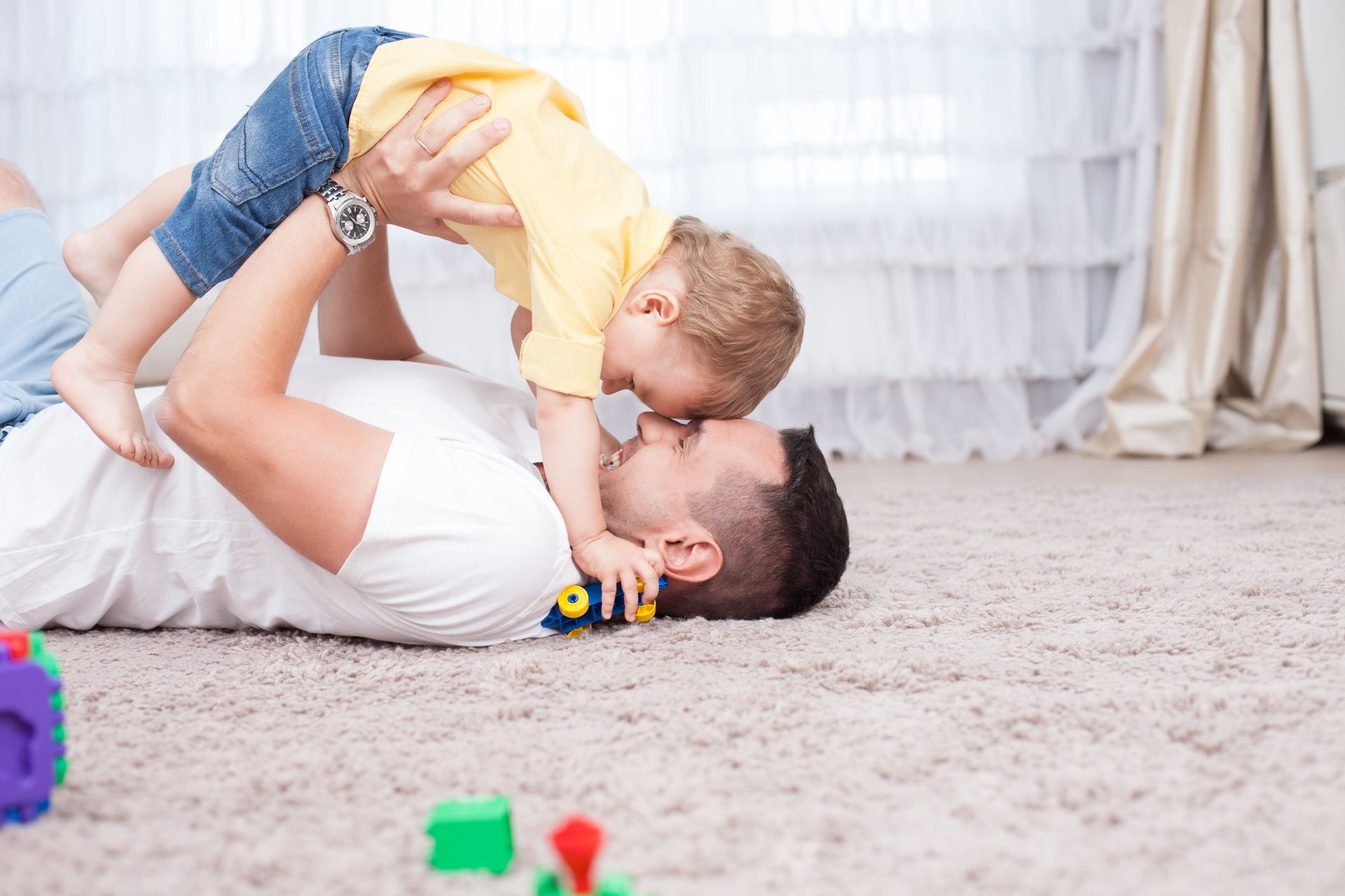 Person installing gray carpet tiles over a blue underlayment. Two children play on the floor, one lying down while the other leans over them with a toy car.