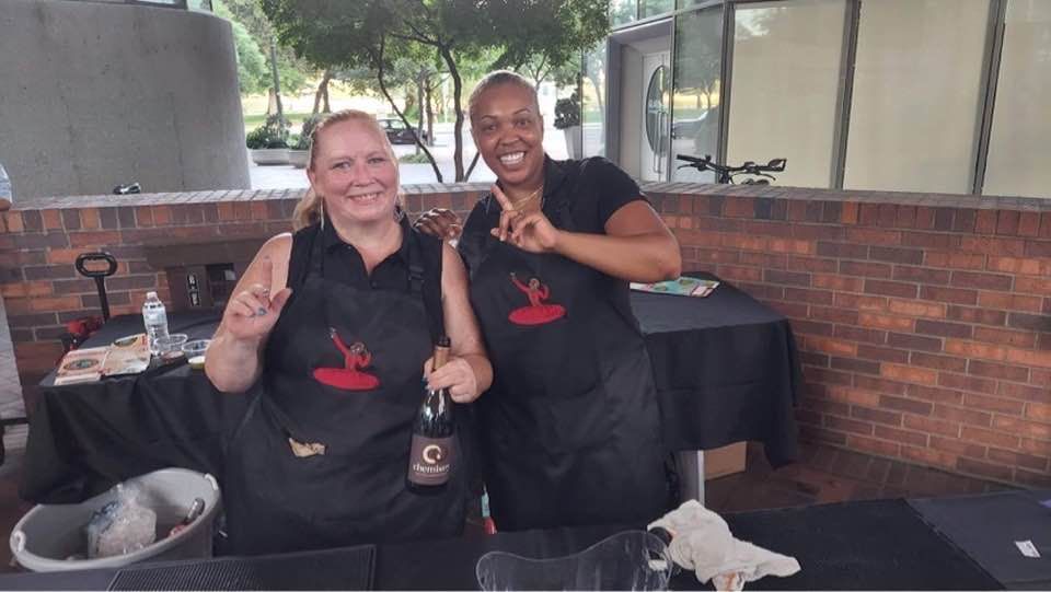 Two women wearing aprons are standing next to each other in front of a table.