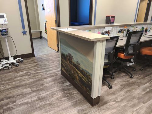Reception desk in a medical office, with a landscape painting on the front. Light wood-look flooring.