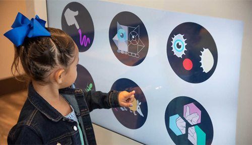 Girl pointing at interactive touch screen with geometric shapes.