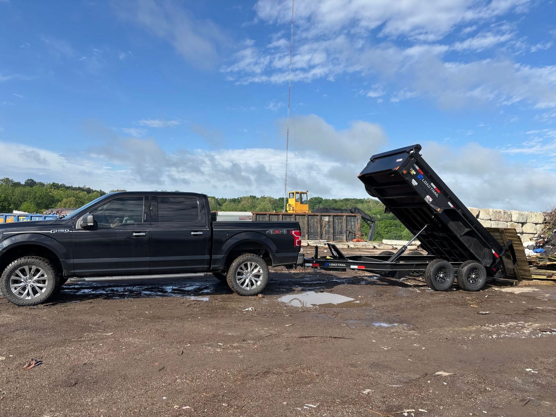 Black pickup truck towing a raised black dump trailer on a gravel lot under a blue sky