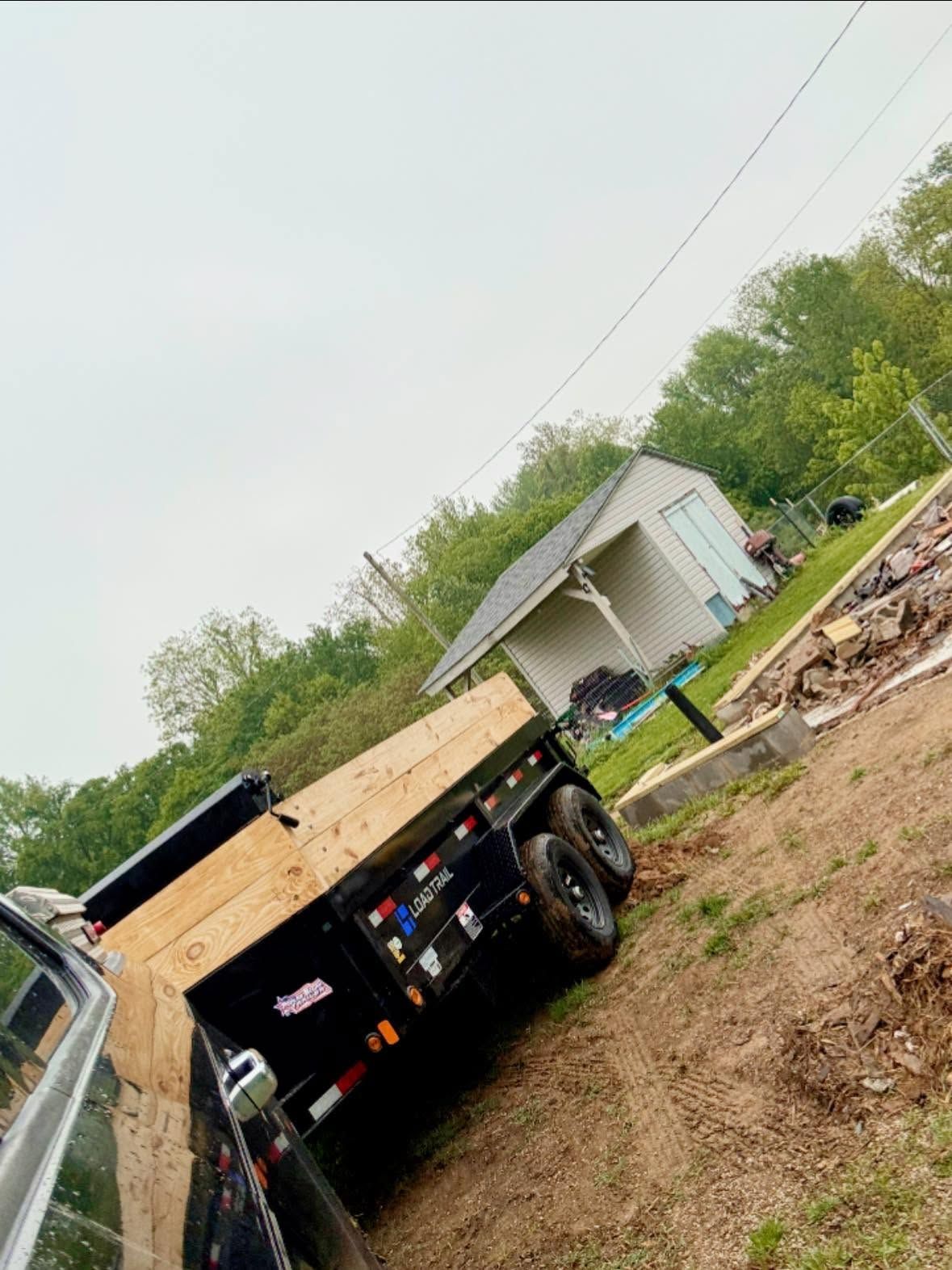 Dump truck and flatbed truck at a muddy rural work site beside a small building.