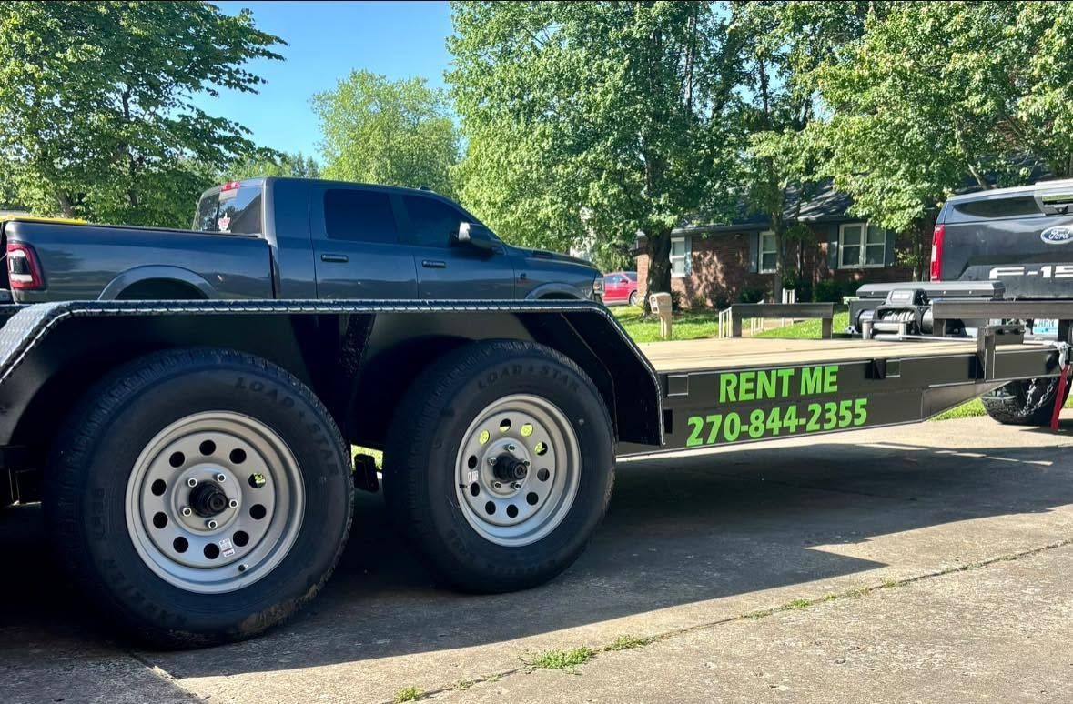 Black dump truck on a flatbed trailer with “RENT ME” sign, parked outdoors by trees.