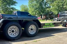 Black dump truck on a flatbed trailer with “RENT ME” sign, parked outdoors by trees.