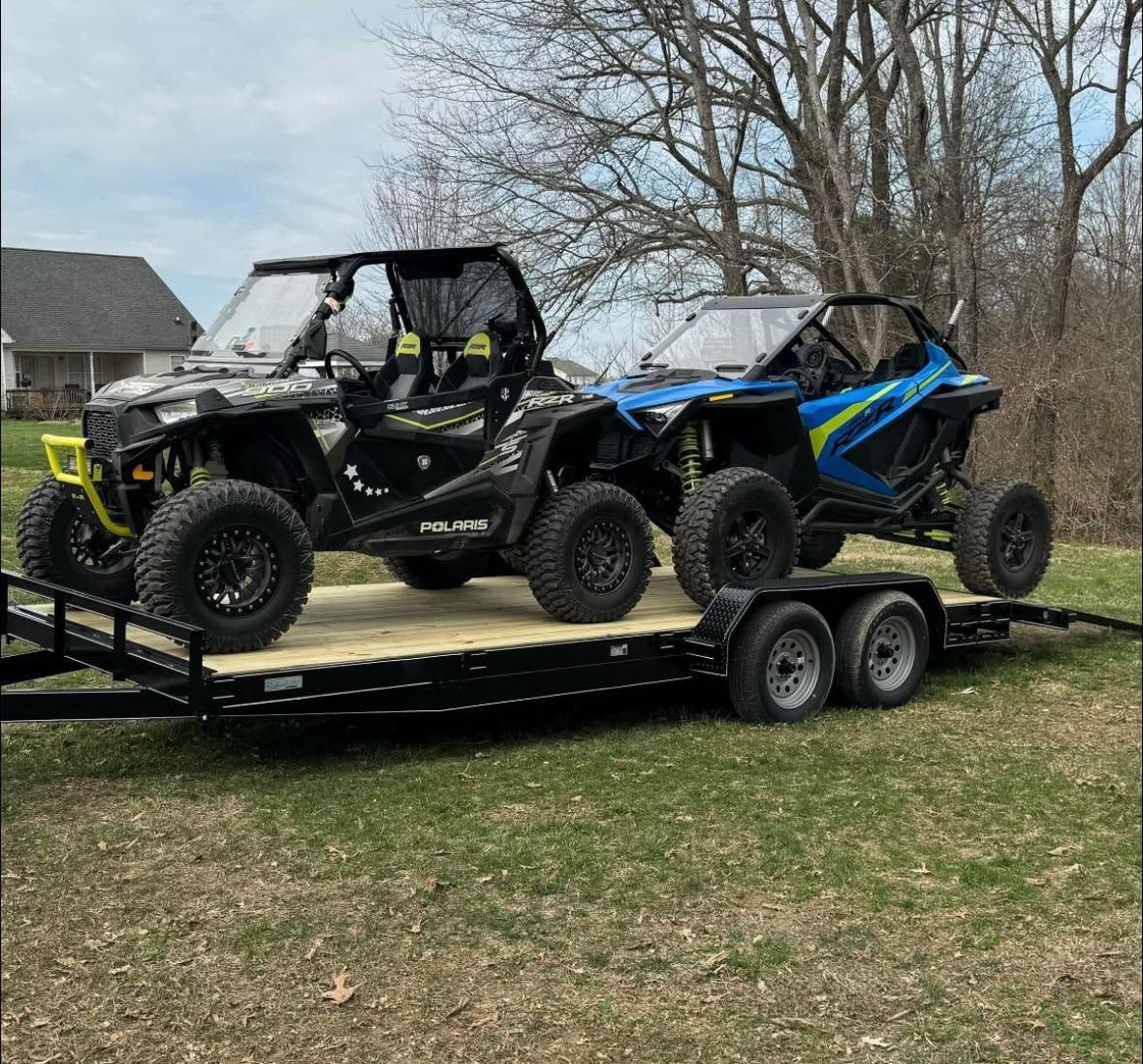 Two off-road UTVs, one black and one blue, parked on a trailer in a grassy field.