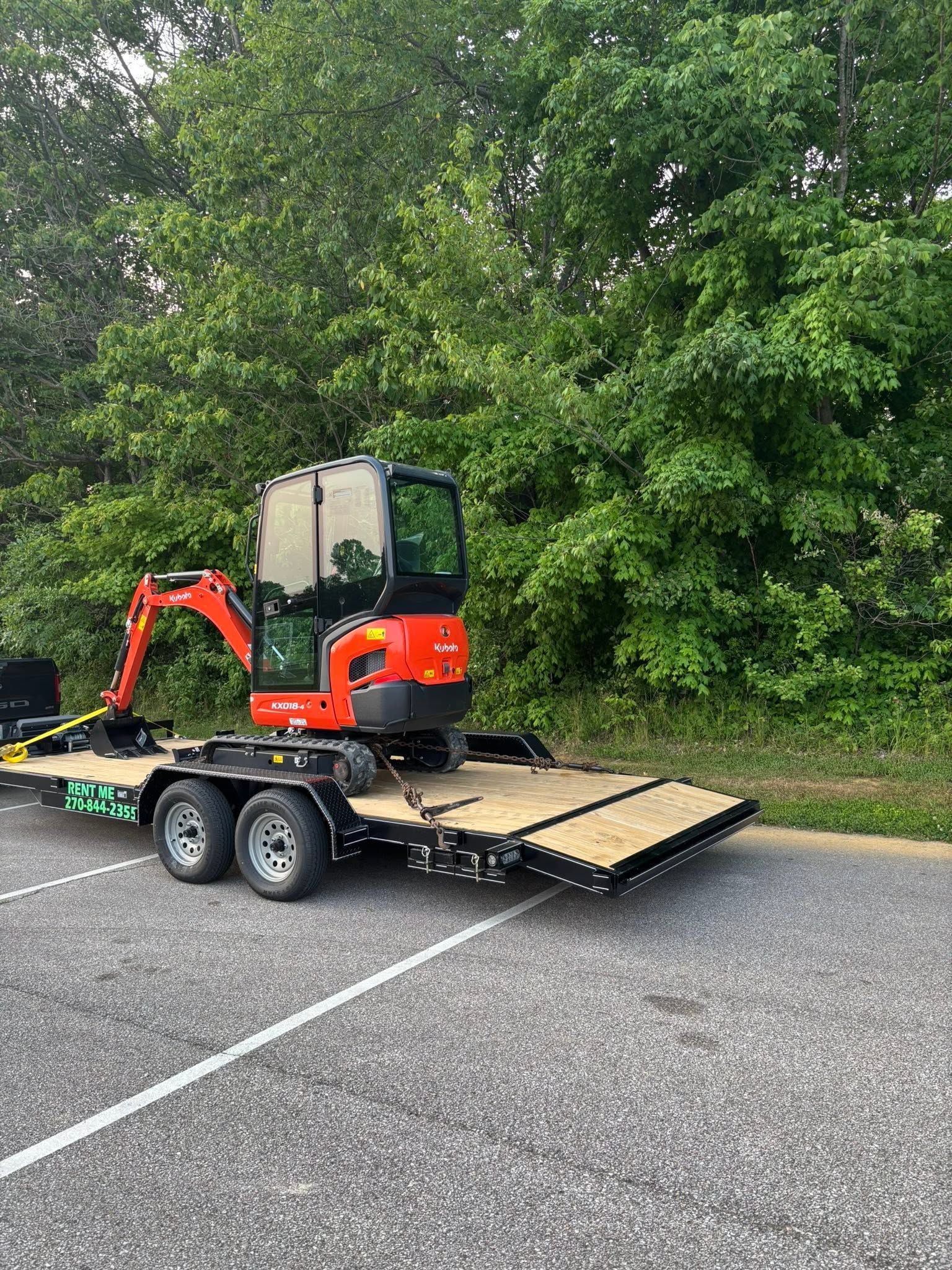 Red mini excavator on a trailer ramp in a parking lot beside trees