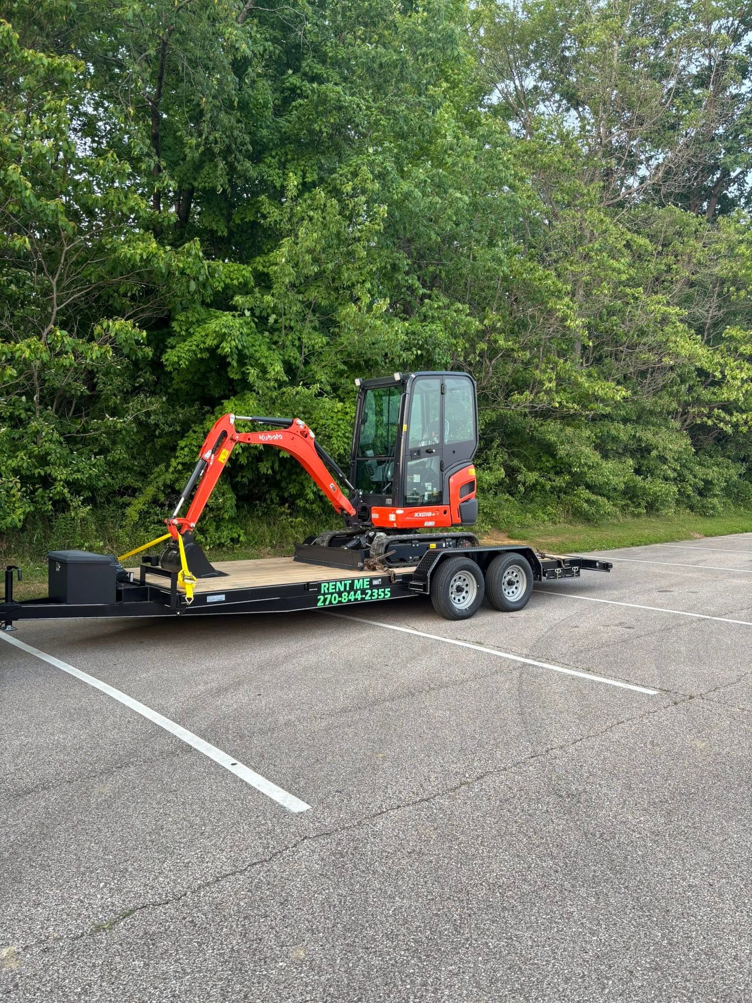 Small orange excavator on a trailer in a gravel lot beside dense green trees.
