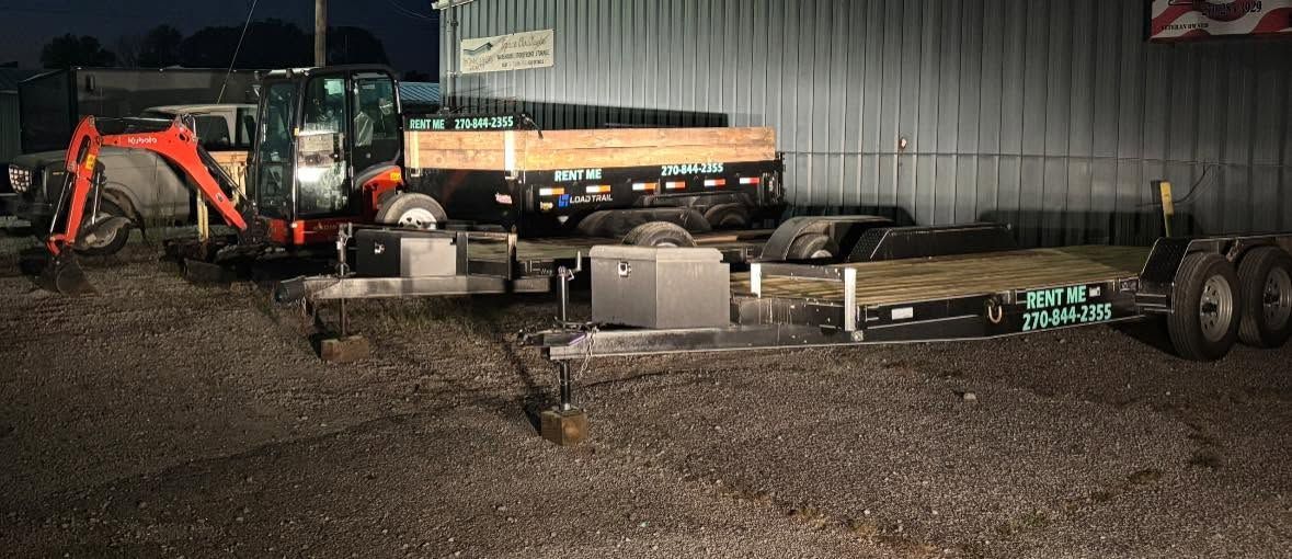 Nighttime farmyard with an orange tractor and a long trailer parked on gravel beside a metal building.