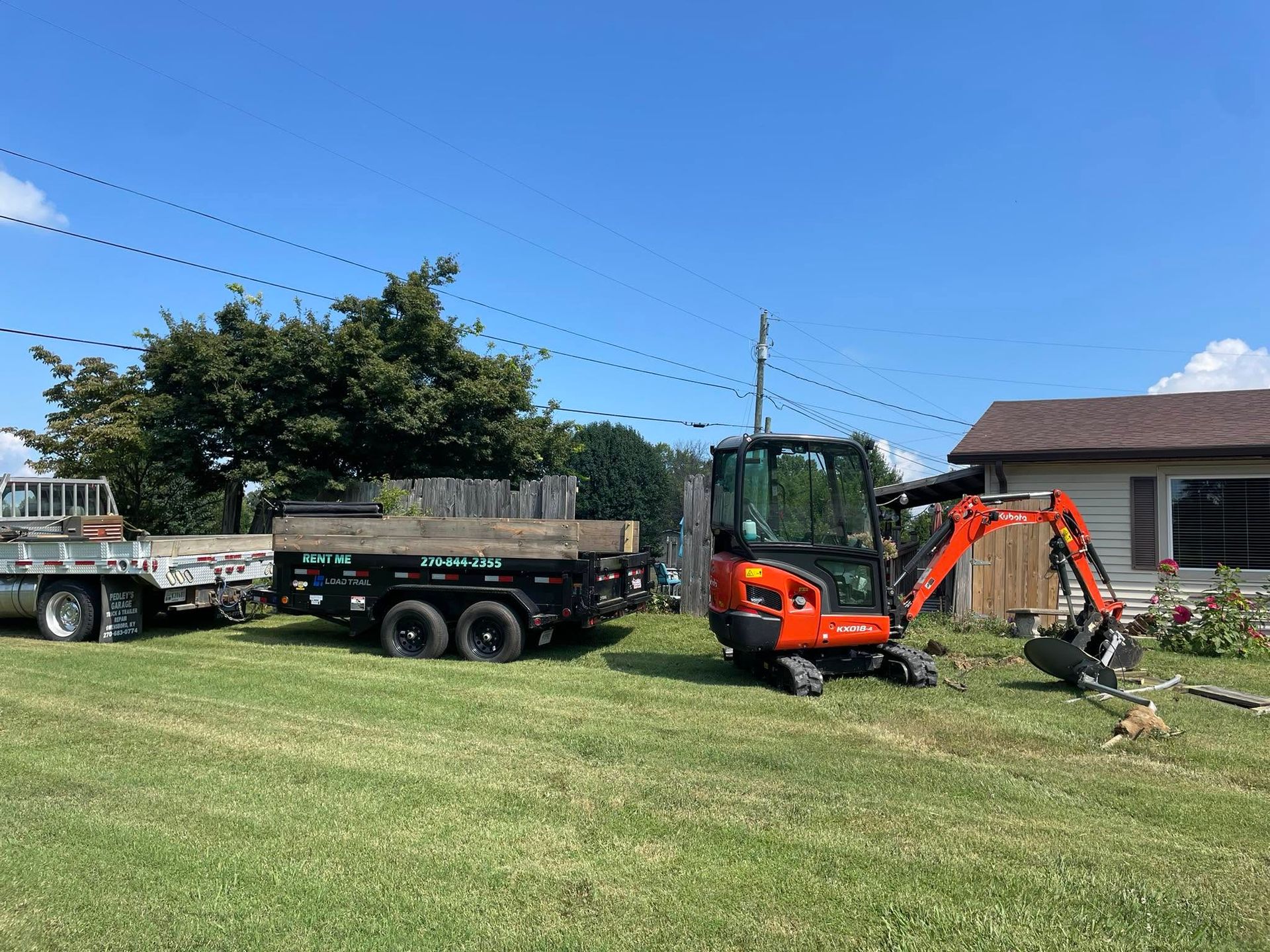 Orange backhoe digging near a black dump trailer on a grassy yard under a clear blue sky