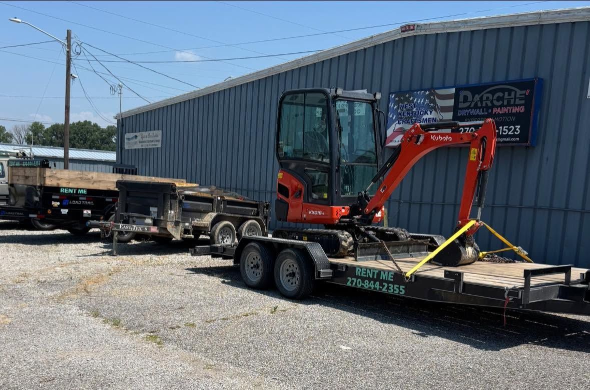 Red excavator on a flatbed trailer beside a blue warehouse on a sunny day.