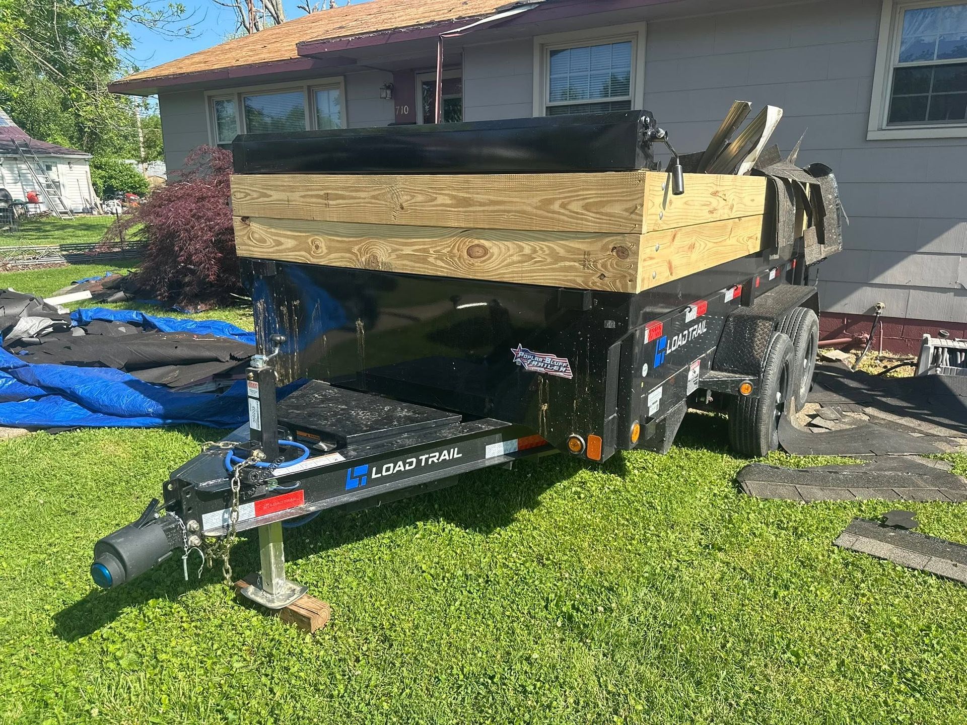 Black utility trailer with wooden cargo sides parked on a lawn beside a house.