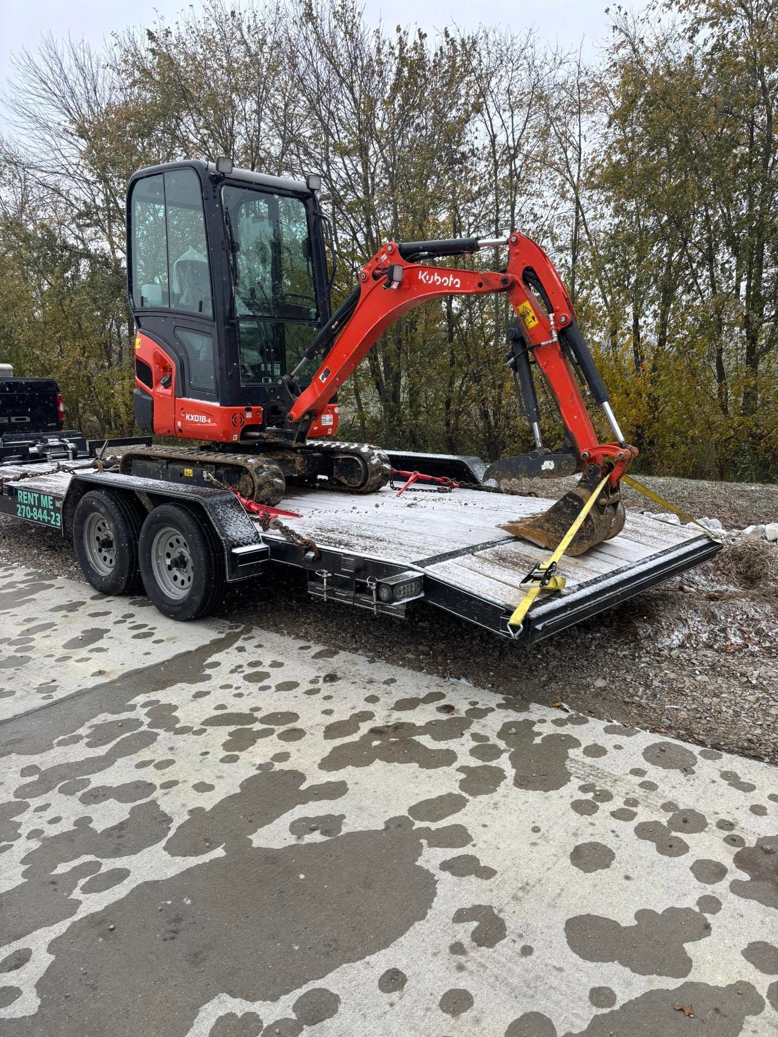 Red mini excavator on a flatbed trailer in a snowy lot, with trees in the background.