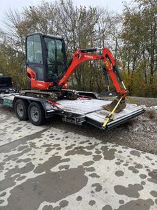 Red mini excavator on a flatbed trailer in a snowy lot, with trees in the background.