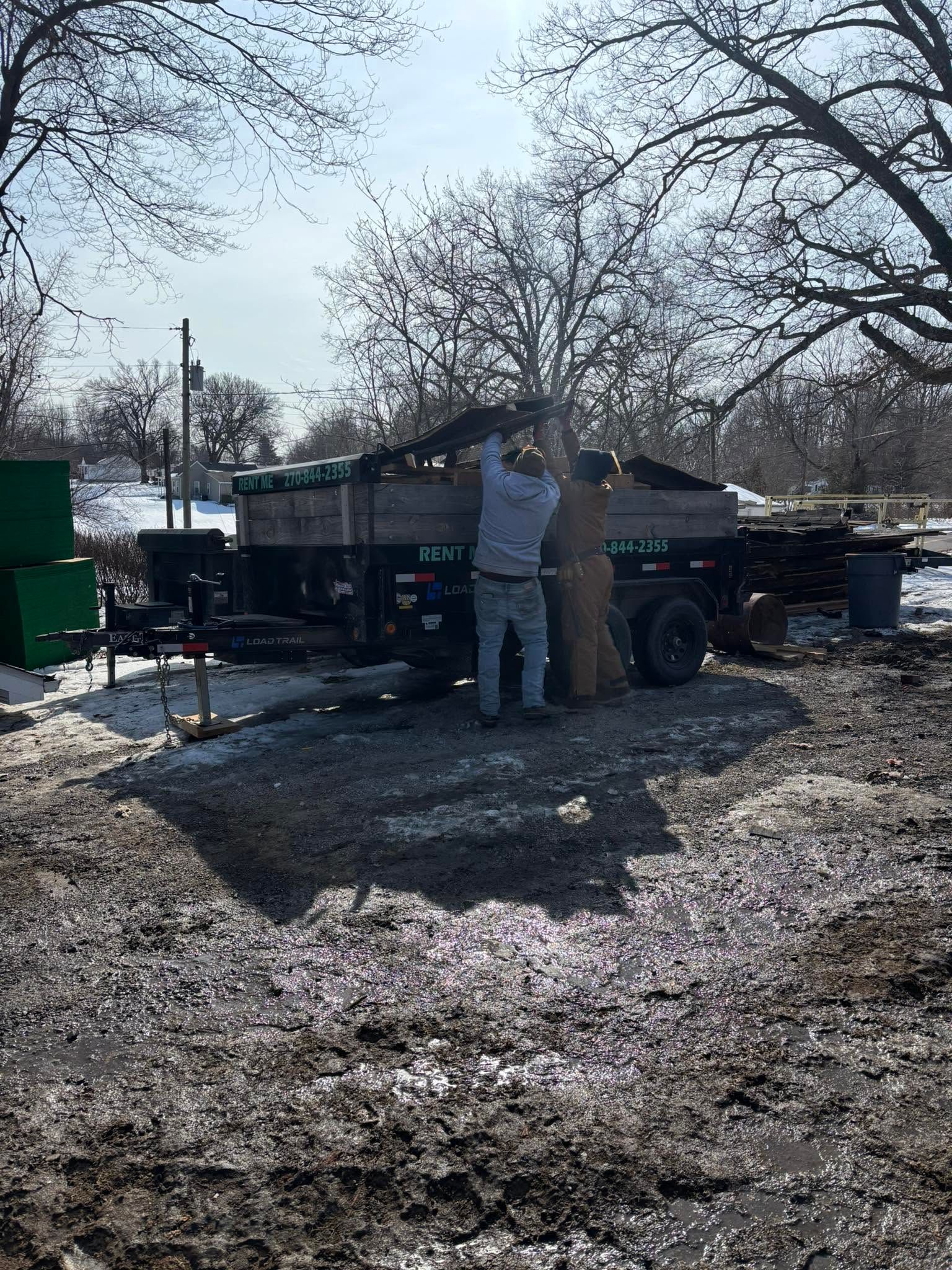 Person loading items into a pickup truck in a muddy yard with bare trees.