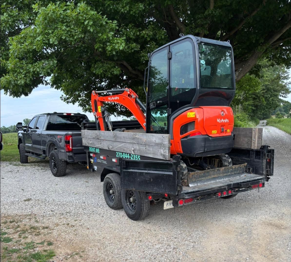 Black pickup towing a trailer with a compact orange excavator on a gravel lot under trees