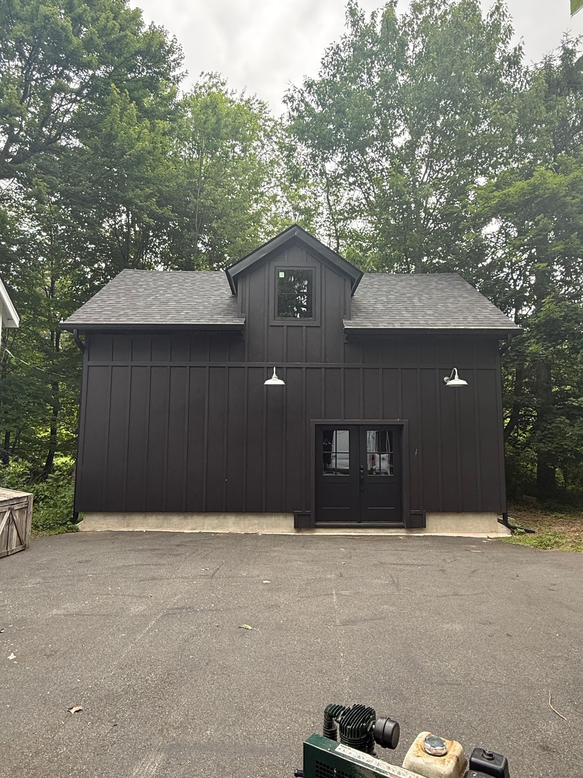 Dark barn-style building with a central door and two lamps, surrounded by trees and gravel.