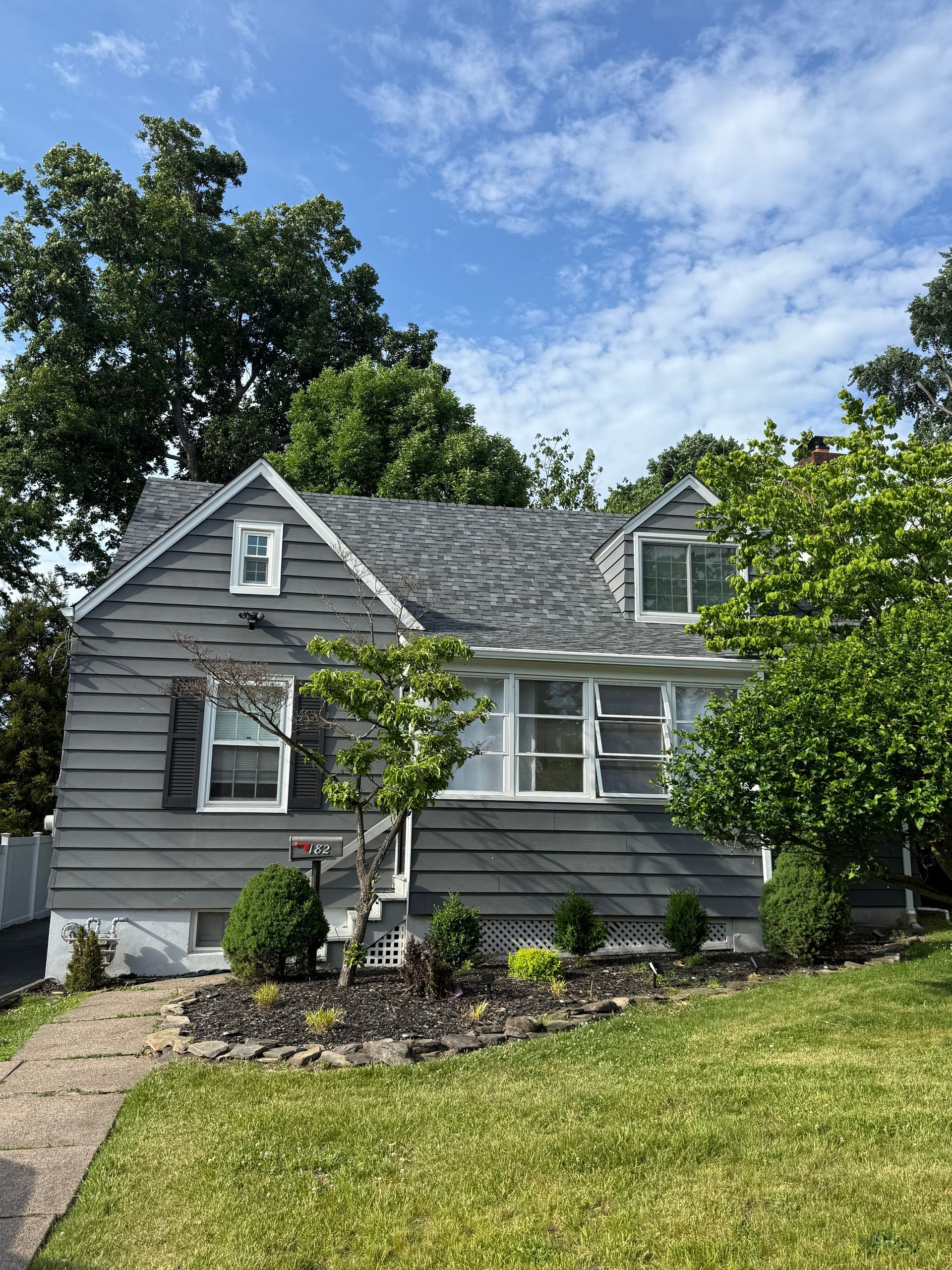 Gray house with white trim, front garden, and green lawn under a partly cloudy sky