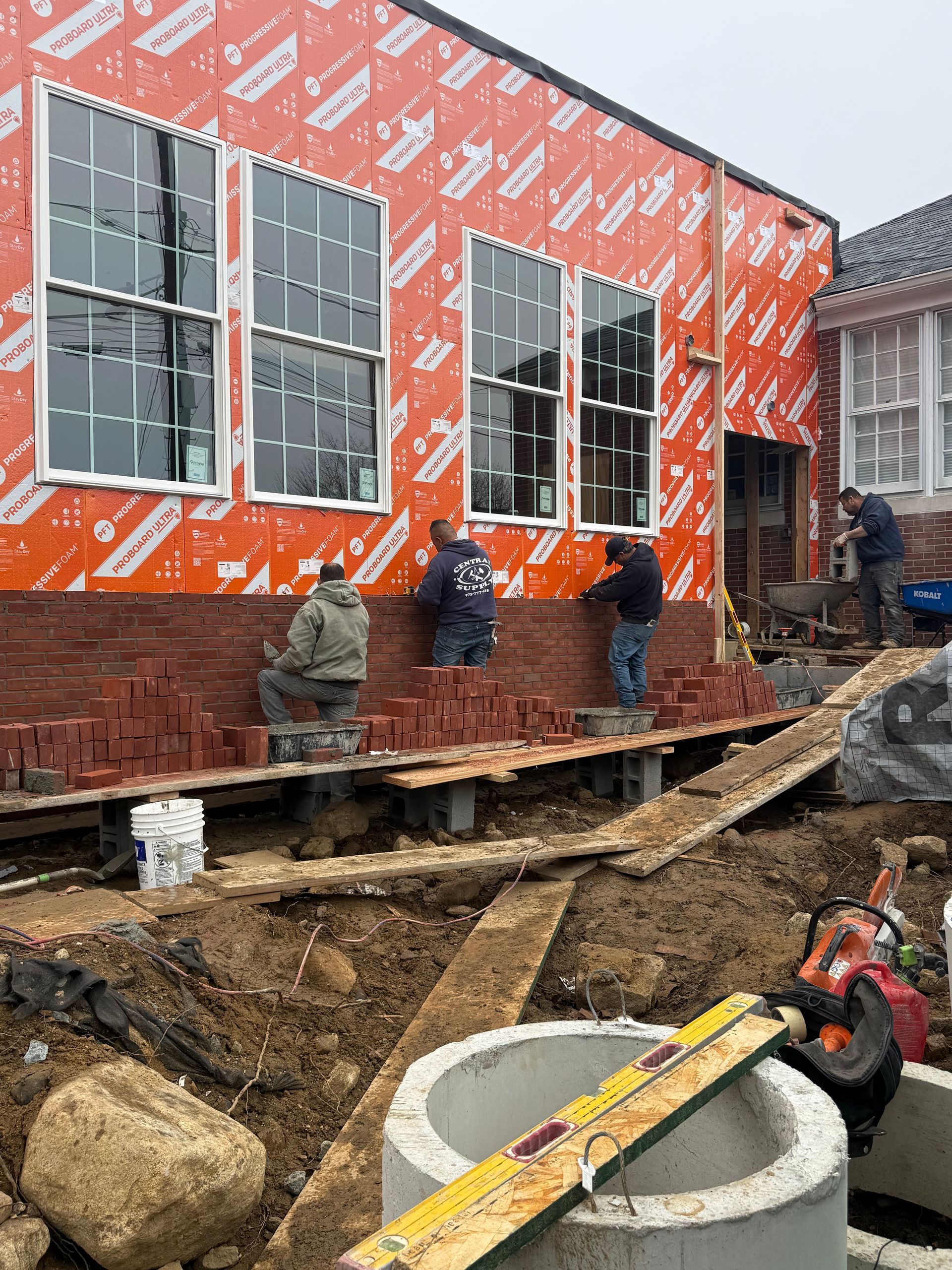 Construction crew working outside a house wrapped in orange housewrap, with a large concrete pipe in the foreground.