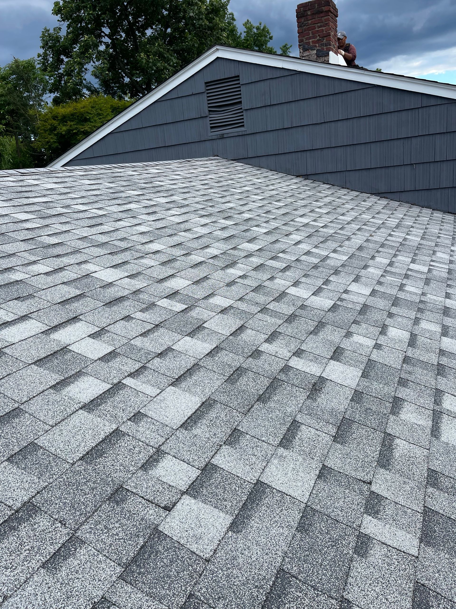 Gray shingled roof on a house under a blue sky, with trees and a chimney in the background