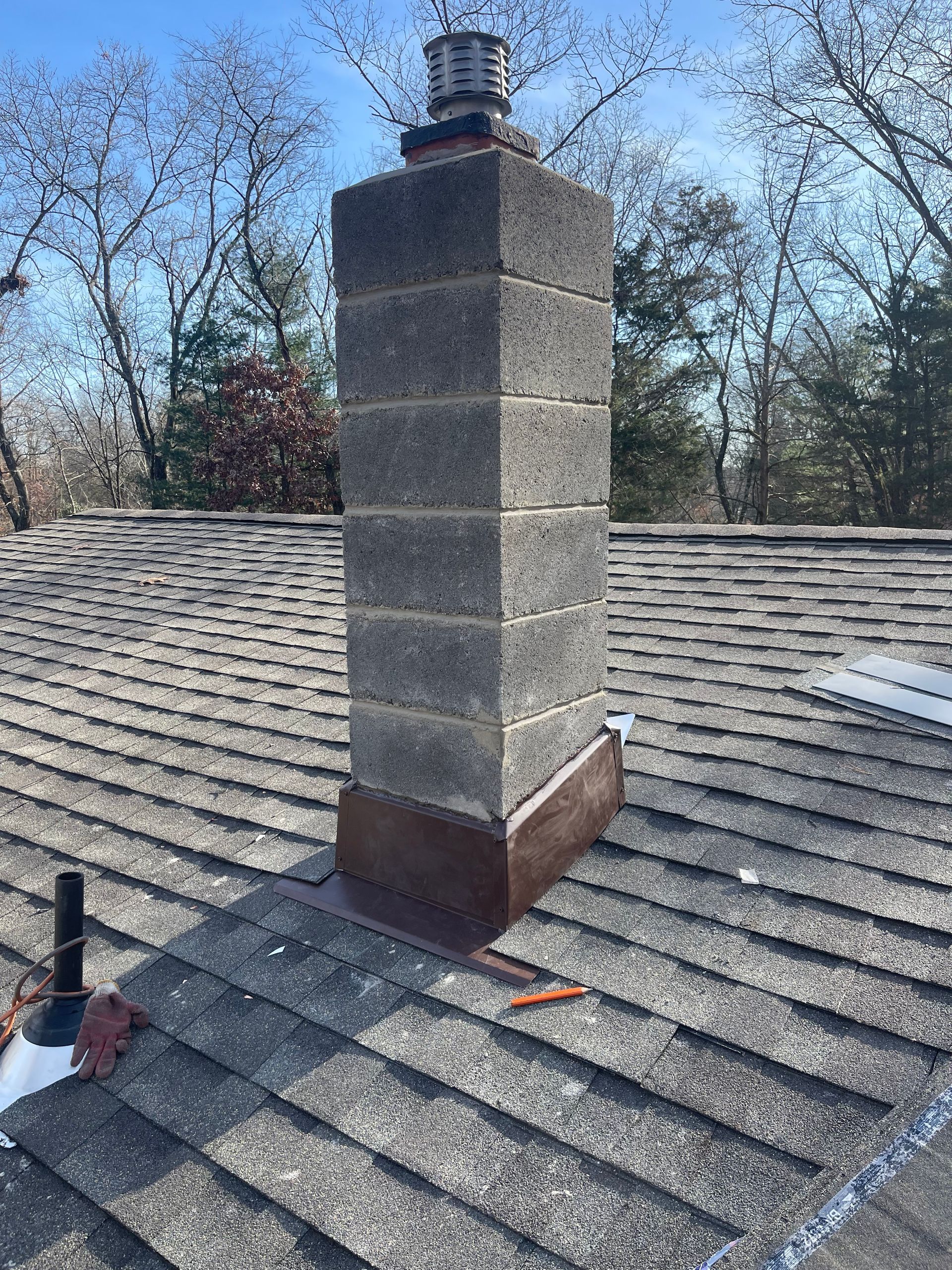 Rooftop chimney with a gray cinder block flue and metal cap on shingled roof, trees in the background