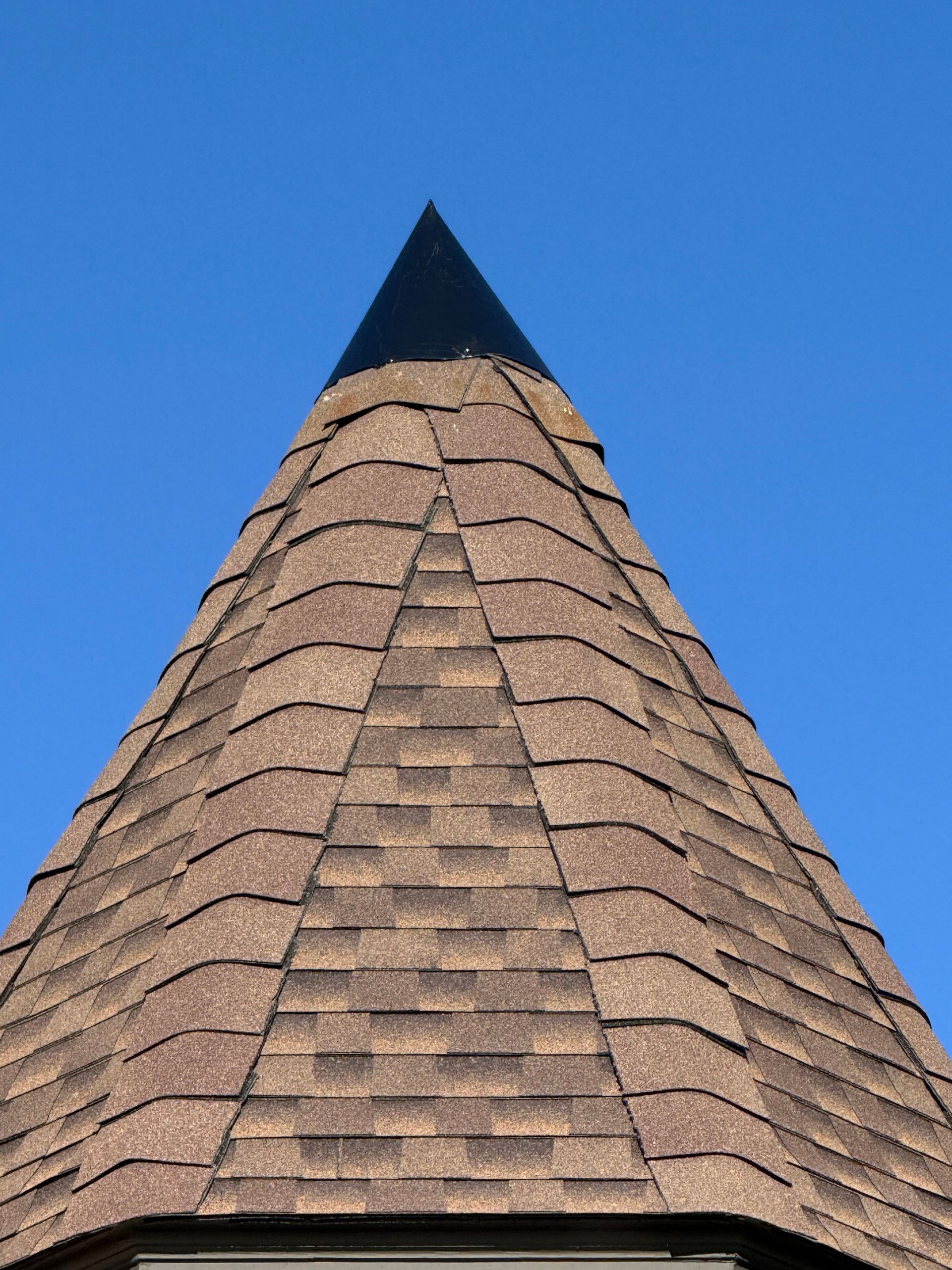 Conical shingled roof with a dark pointed cap against a clear blue sky